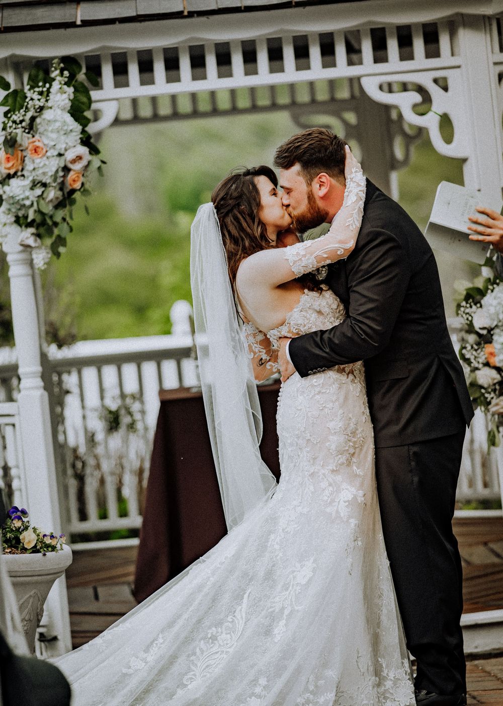Romantic wedding ceremony kiss in a decorated white gazebo with flowers and lace dress details.