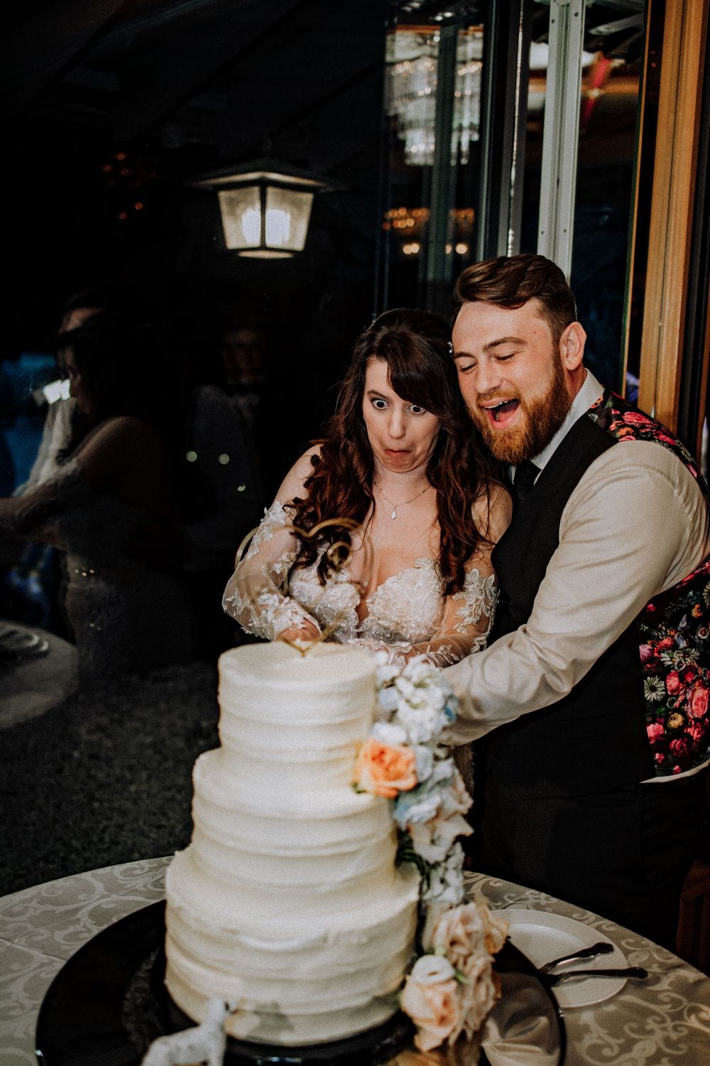 A couple stands together cutting their elegant three-tiered white wedding cake decorated with fresh flowers.