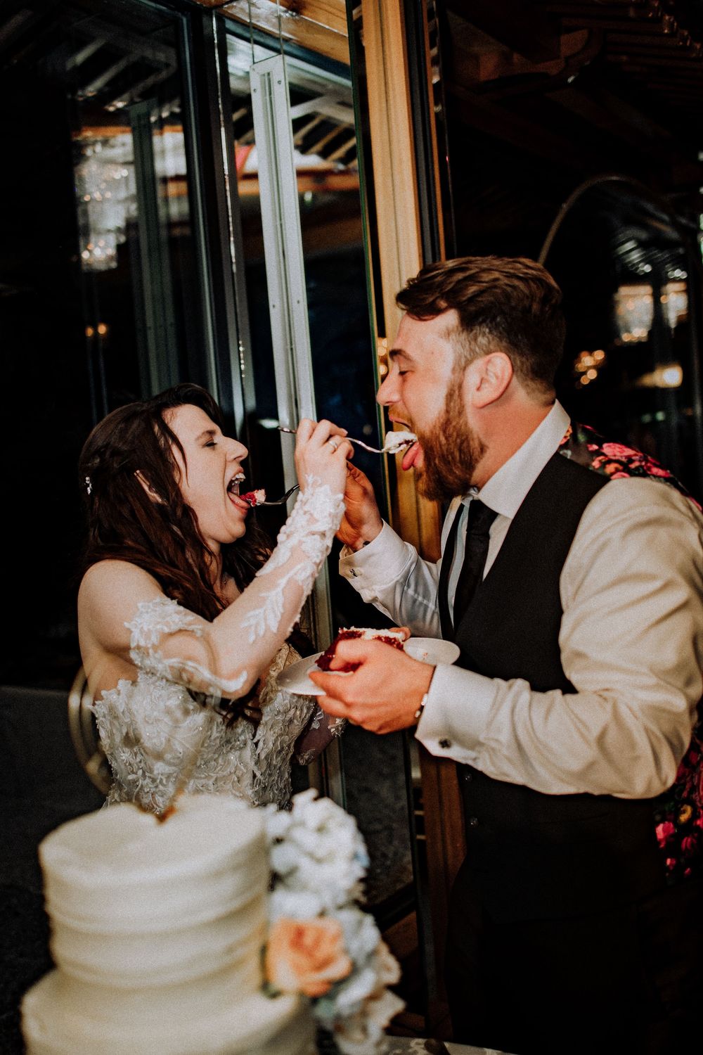 Newlywed couple shares a playful moment feeding each other wedding cake beside their decorated white wedding cake.