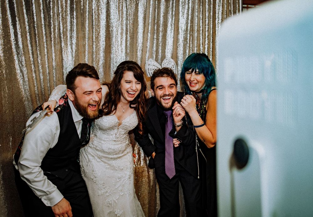 Happy wedding party members pose together inside a photo booth with a silver sequin backdrop.