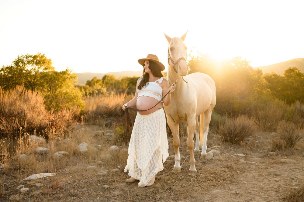 woman stands with white horse in two piece white lace dress in sunlight at Ojai Valley Trail riding during her maternity photoshoot in Ojai
