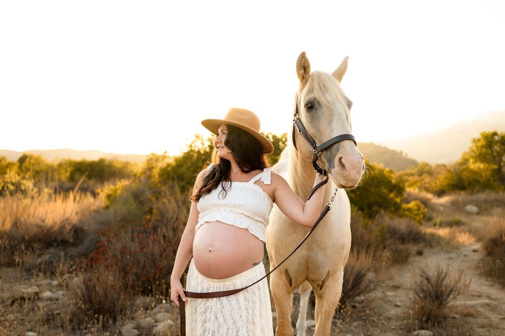 woman poses with white horse during sunset in cowgirl outfit at Ojai Valley Trail riding during her maternity photoshoot