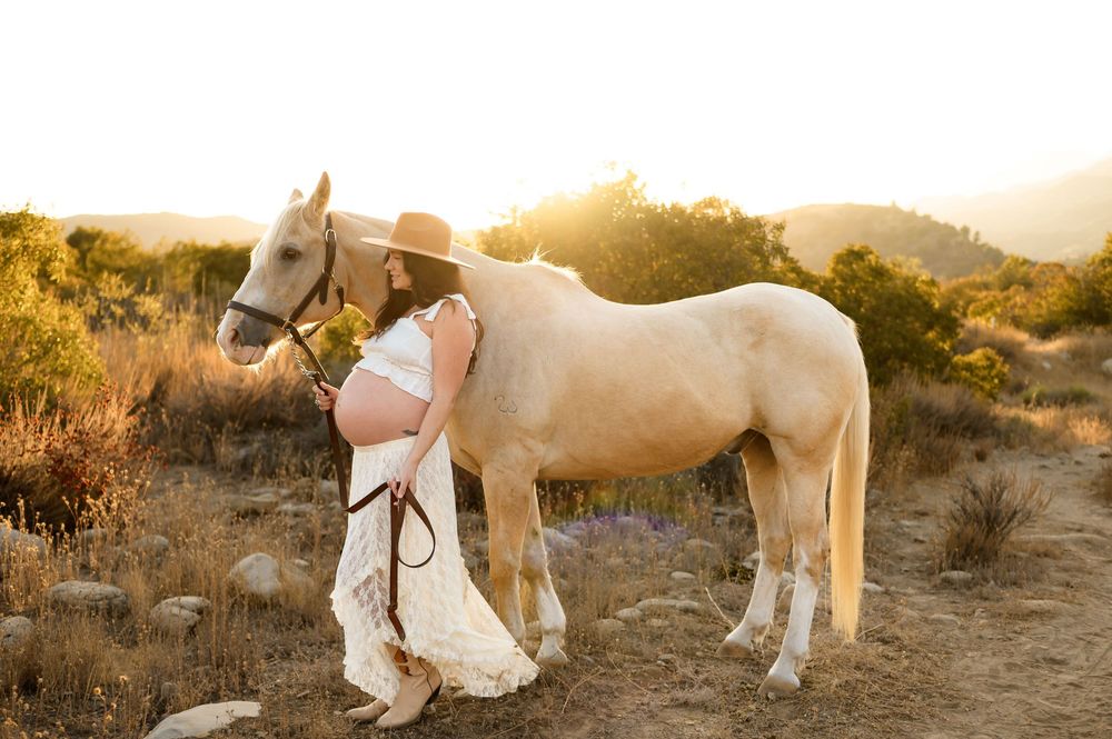 woman leans against white horse in two piece white lace dress and cowboy boots at Ojai Valley Trail riding during her maternity photoshoot
