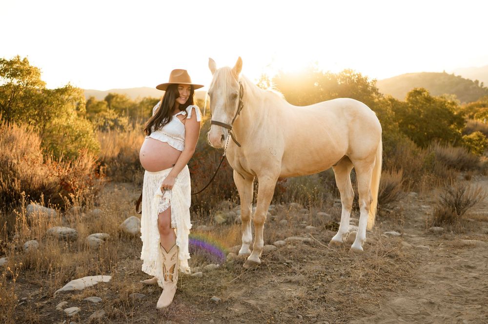woman poses with white horse during sunset in cowgirl outfit at Ojai Valley Trail riding during her maternity photoshoot