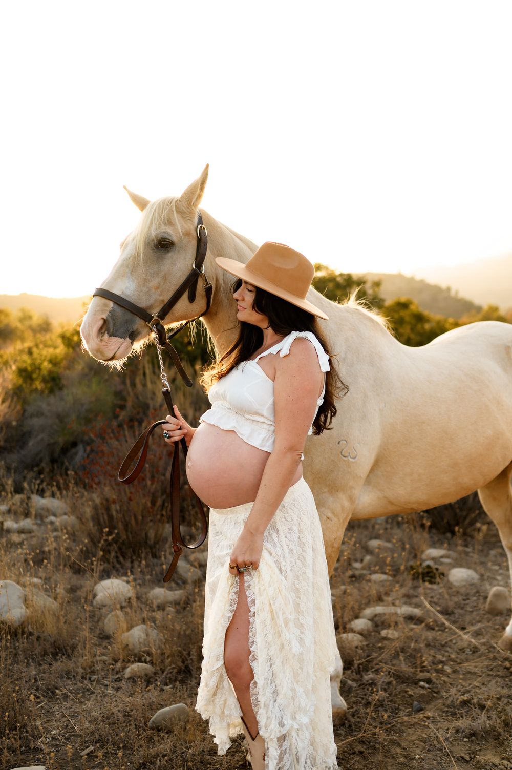 woman stands with white horse in white lace dress during her maternity photoshoot in Ojai river preserve at Ojai Valley Trial Riding Company