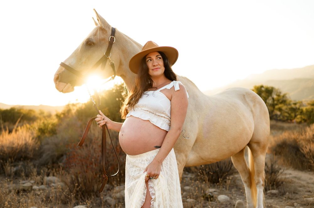 woman and horse pose in sunlight in cowgirl outfit at Ojai Valley Trail riding during her maternity photoshoot on Ojai babymoon