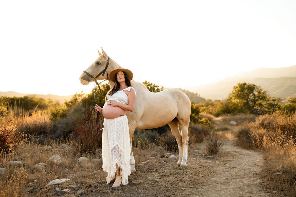 woman stands with white horse in white lace dress & cowboy boots during her maternity photoshoot at Ojai Valley Trial Riding Company