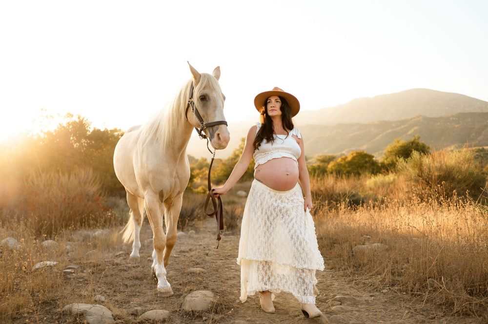 woman walks with horse into sunset in white lace dress and cowboy hat during her maternity photoshoot at Ojai Valley Trial Riding Company