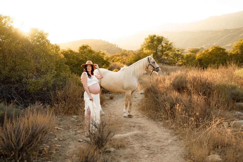woman stands with white horse on trail in front of oak trees at Ojai Valley Trail riding during her maternity photoshoot in Ojai