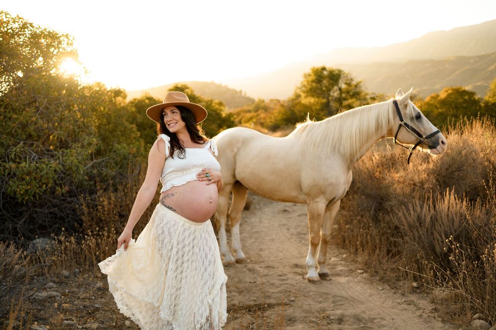 woman in cowboy hat plays with lace skirt in front of white horse during her maternity photoshoot in Ojai river preserve on her babymoon