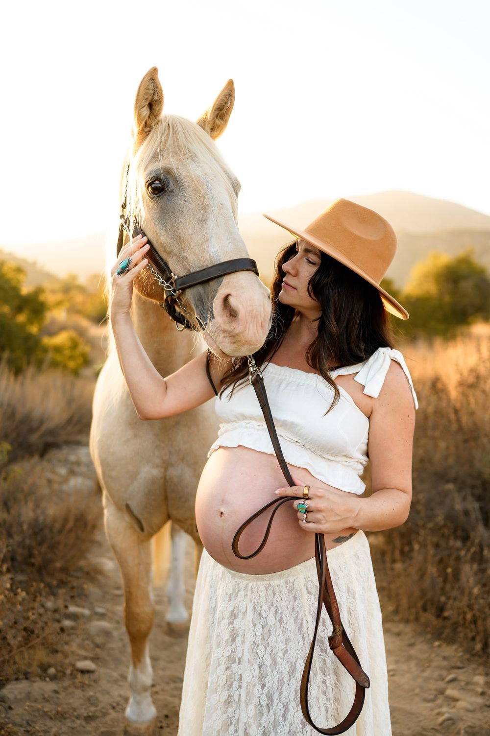 woman poses with white horse holding onto it's face near hers at Ojai Valley Trail riding during her maternity photoshoot