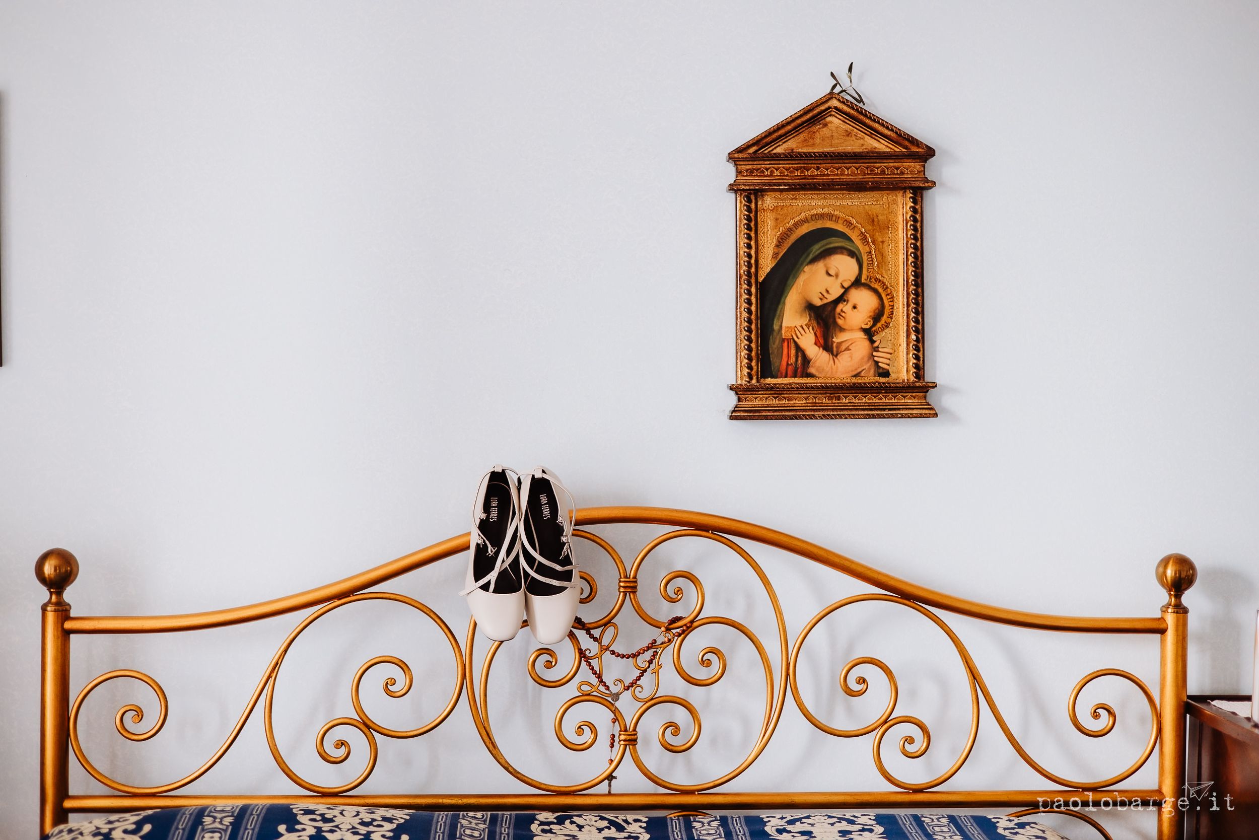 Black ballet shoes hang on a decorative copper-colored metal bed frame beneath a wooden religious wall art piece.