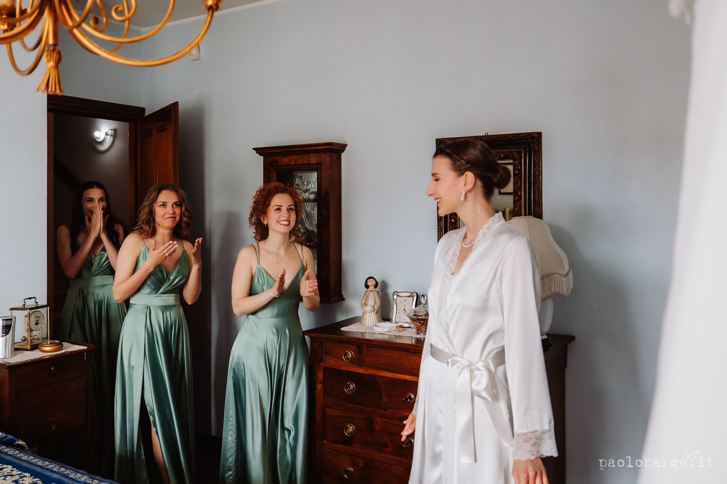 People in matching green dresses look on excitedly while someone in a white robe stands in an elegant dressing room.