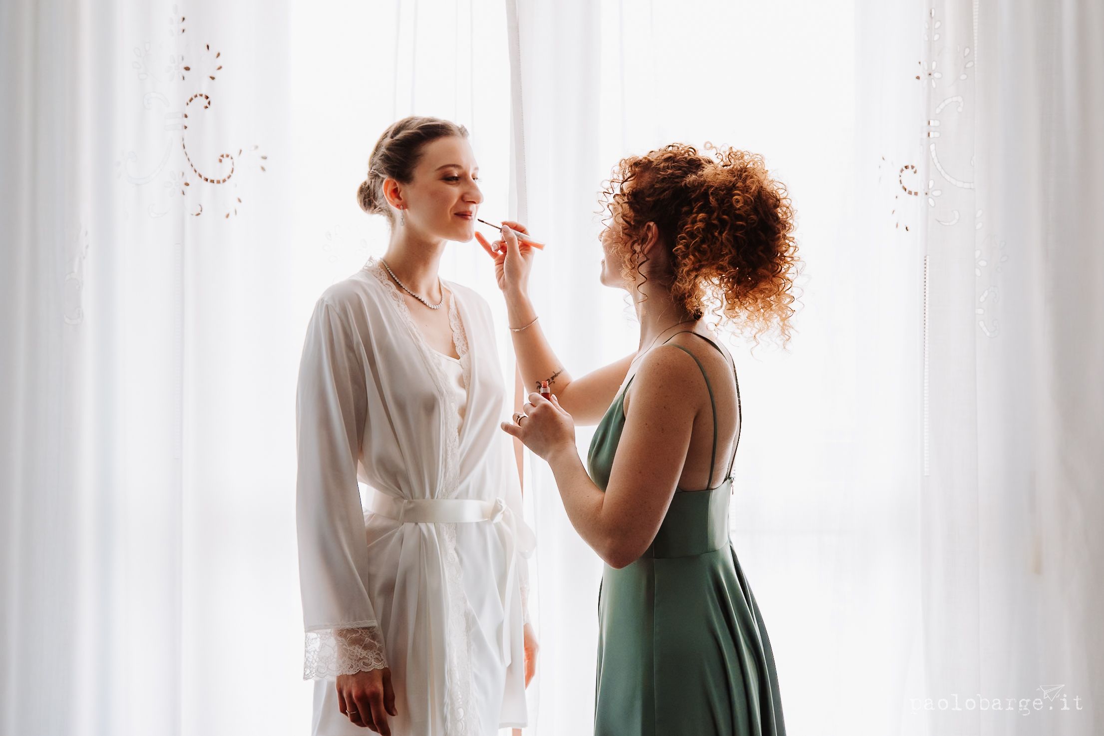 Two people in a sunlit room preparing for a special occasion with one in a white robe and another in a green dress.