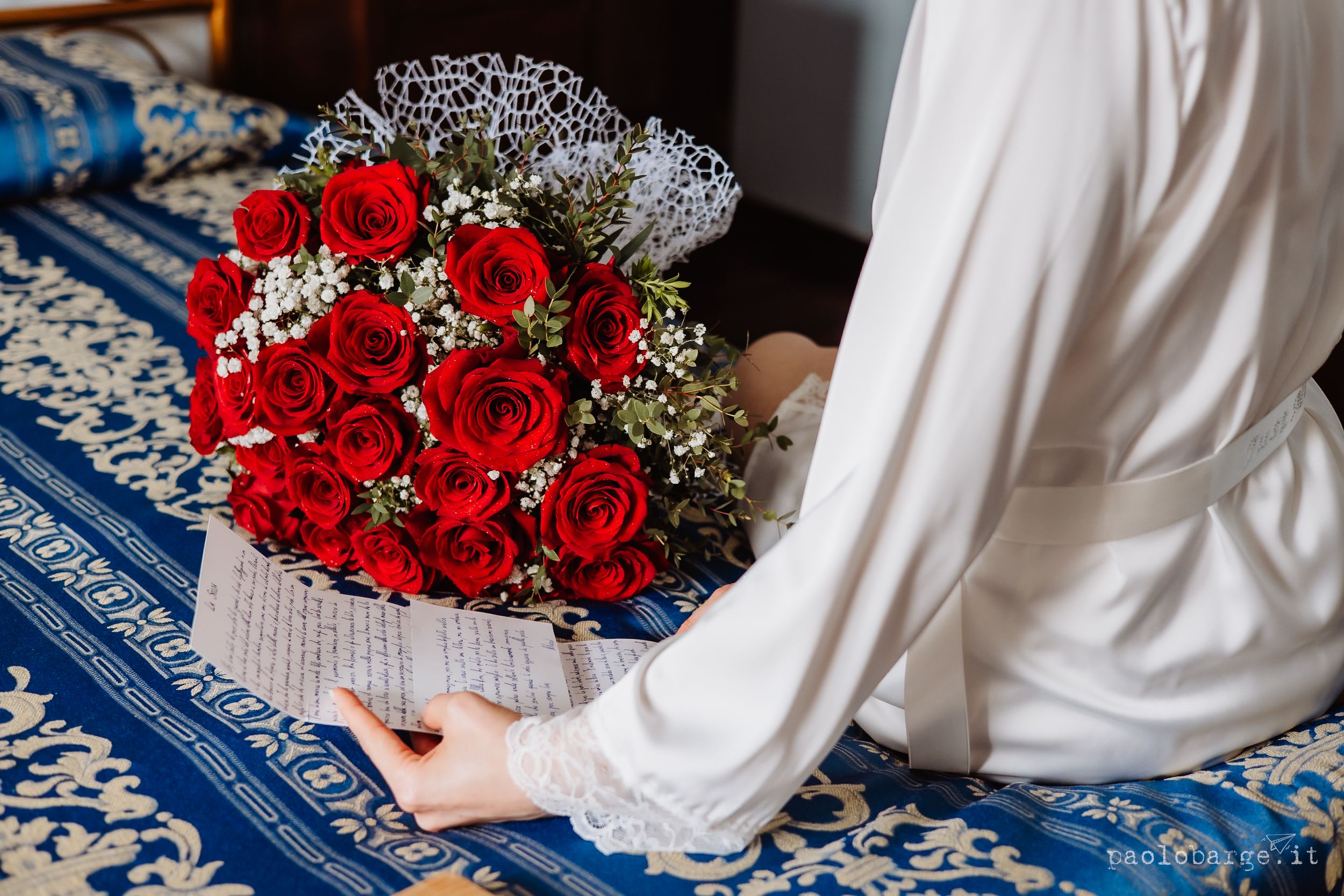 A bridal couple sits on a blue carpet with red roses and white baby's breath flowers scattered around.