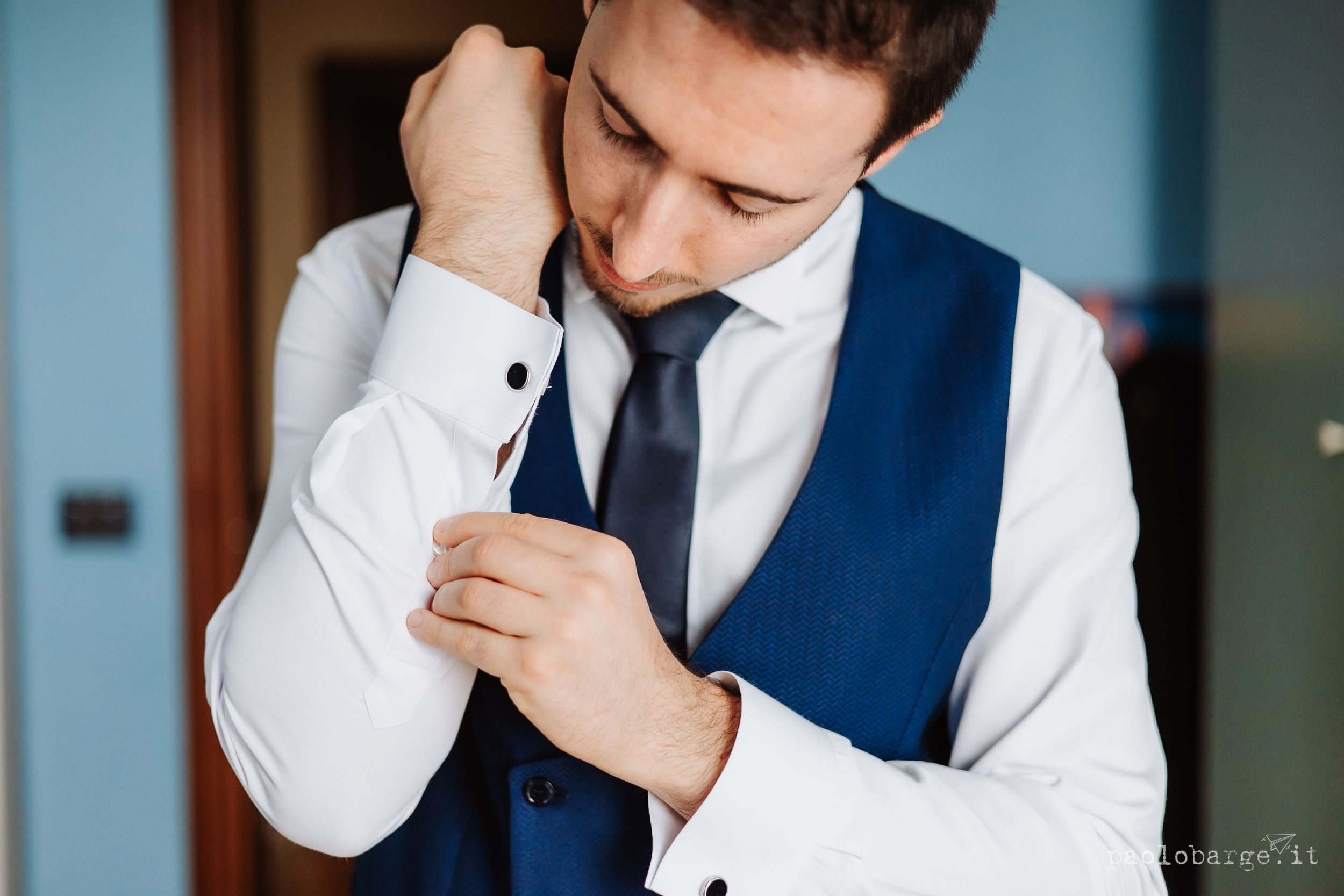 Person in white dress shirt and navy blue vest adjusts their sleeve cuff while getting dressed.