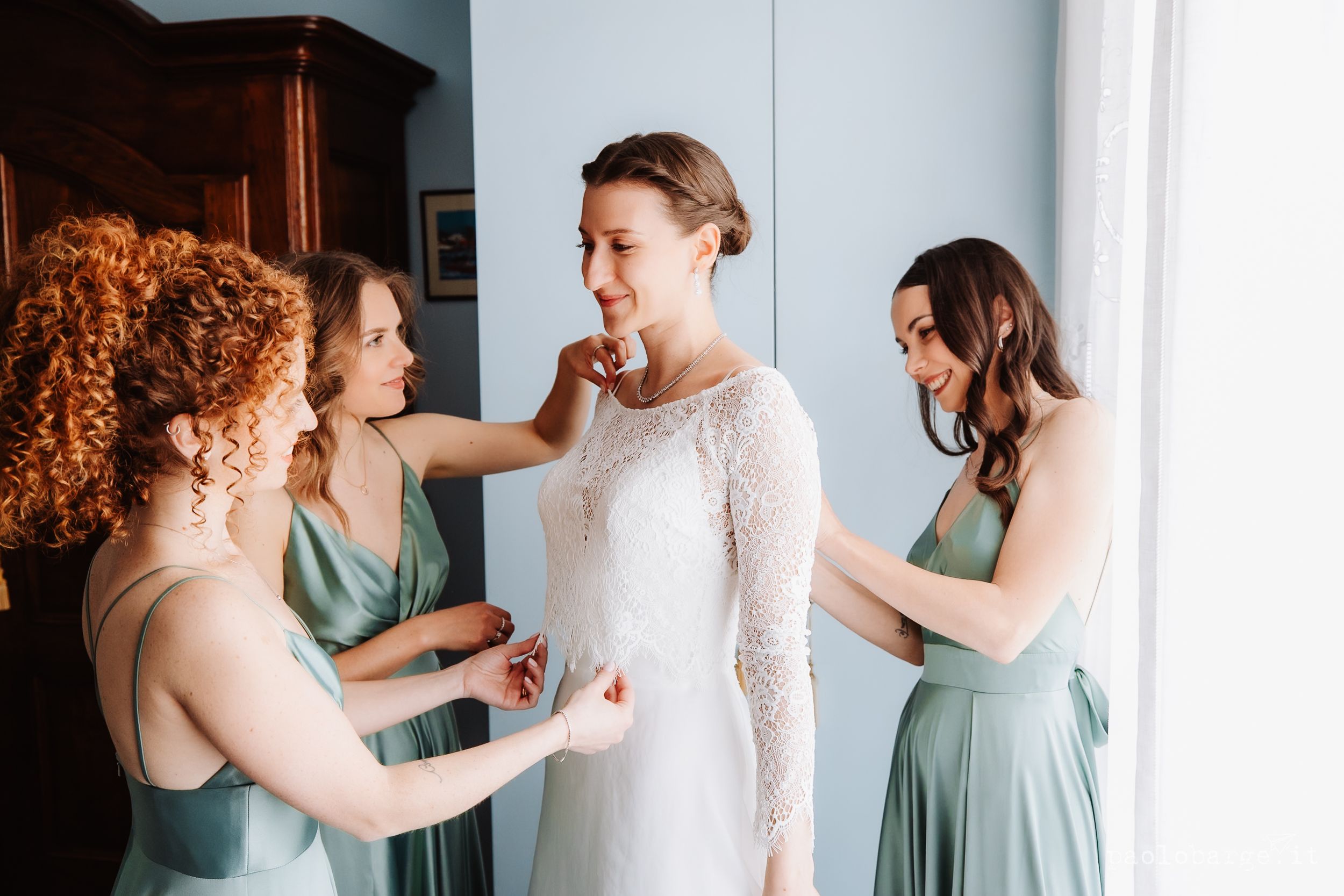 A person in a long white lace dress stands with friends in sage green dresses helping with final adjustments.