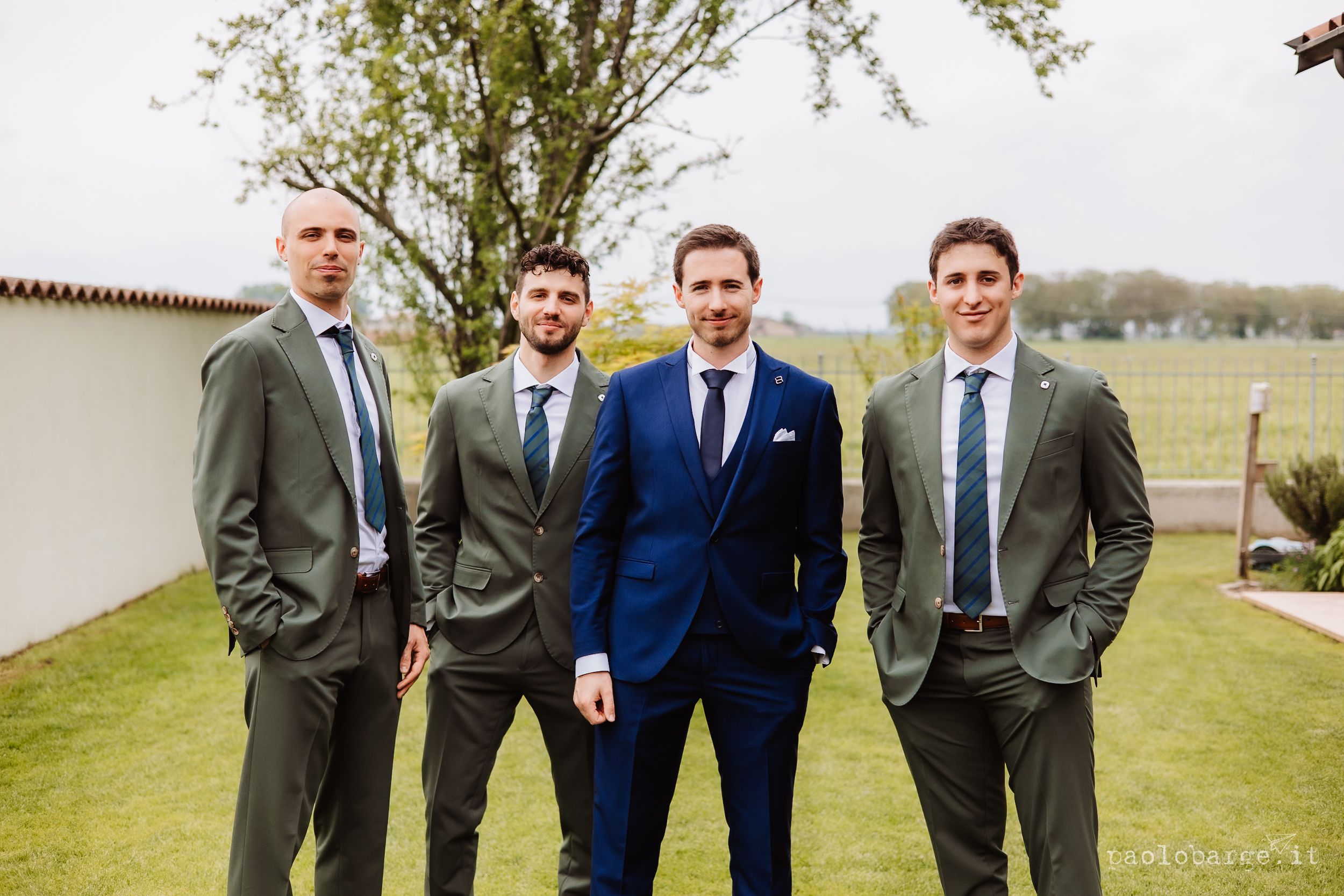 Four men in formal suits standing outdoors on green grass with trees in background during a wedding event.