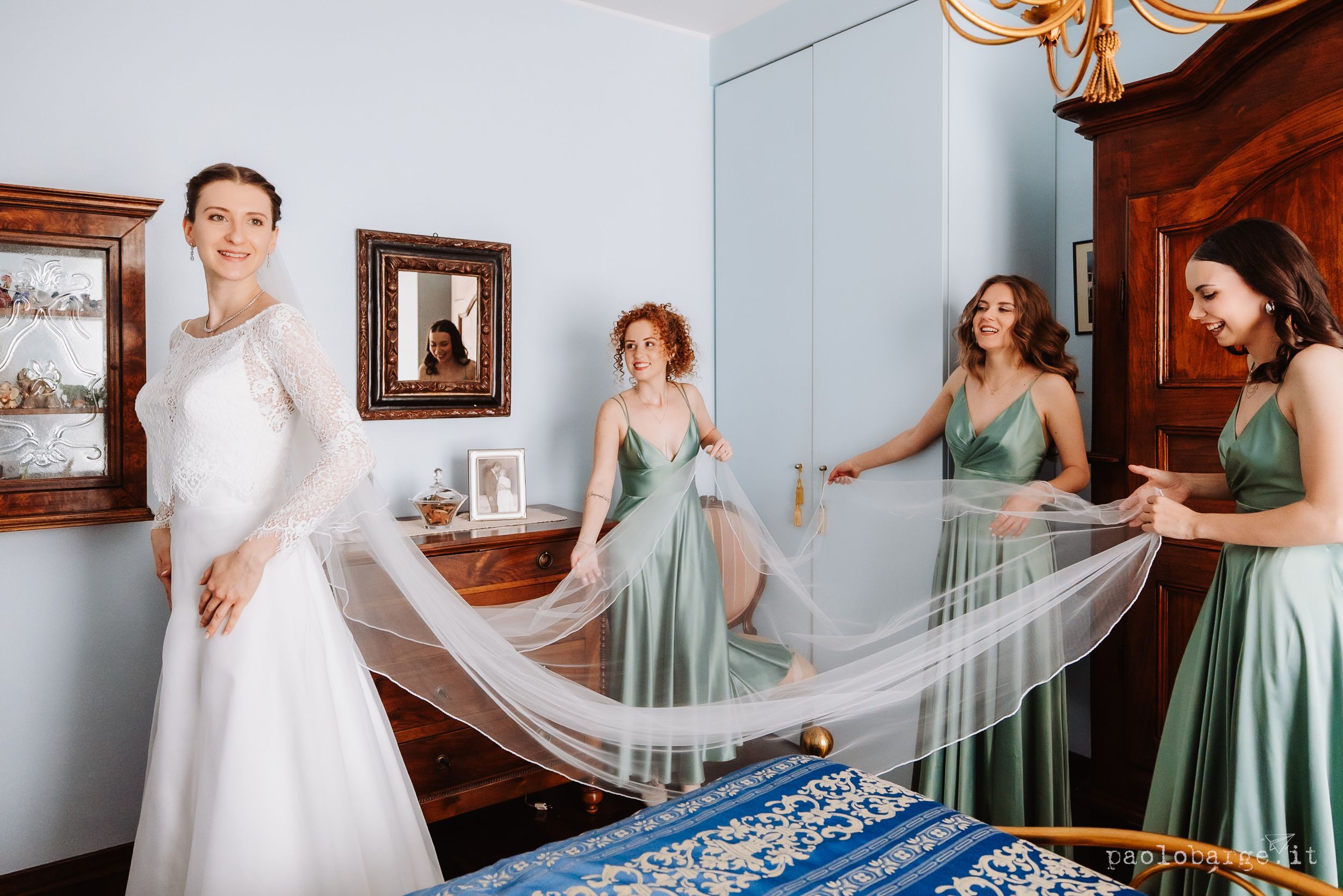 Bride in white gown with bridesmaids in green dresses helping with her veil in an elegant bedroom setting.