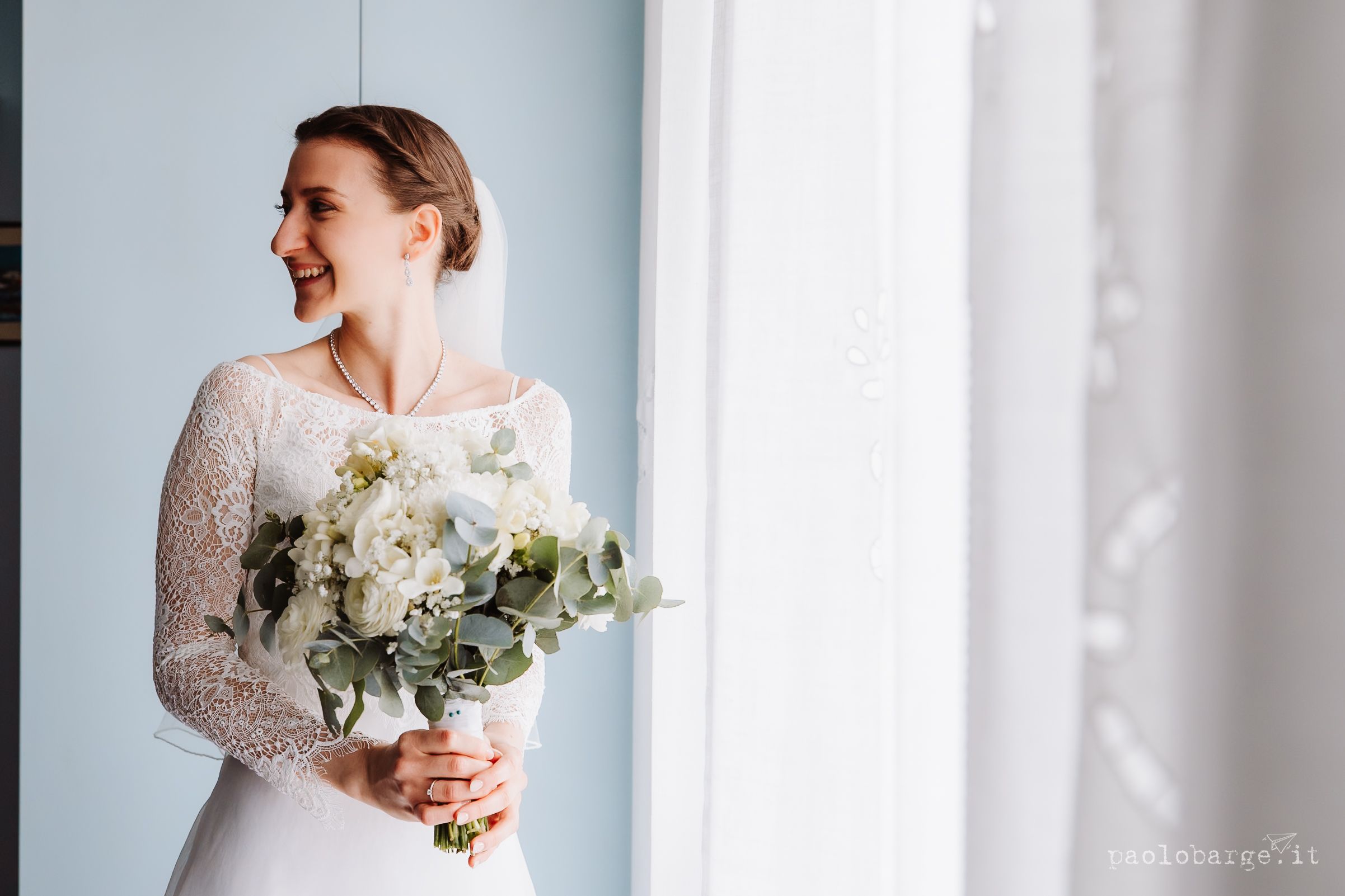 A person in a white lace wedding dress holds an elegant bouquet of white flowers and greenery by a window.