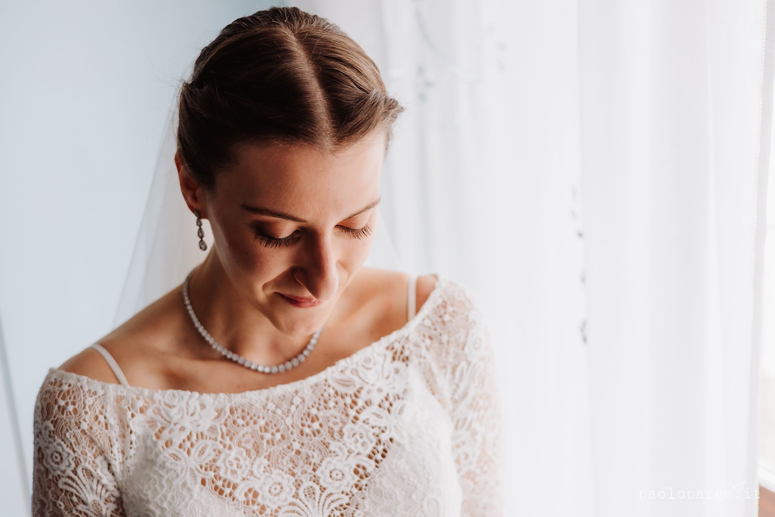 Bride wearing lace dress with scalloped neckline poses gracefully in natural window light during wedding preparations.