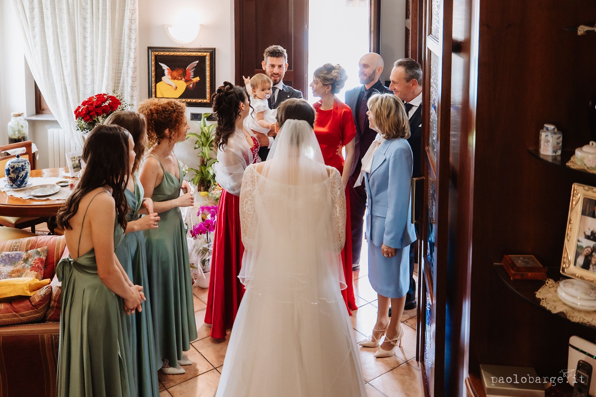 A wedding party gathers in a room as guests in colorful dresses surround the bride in white.