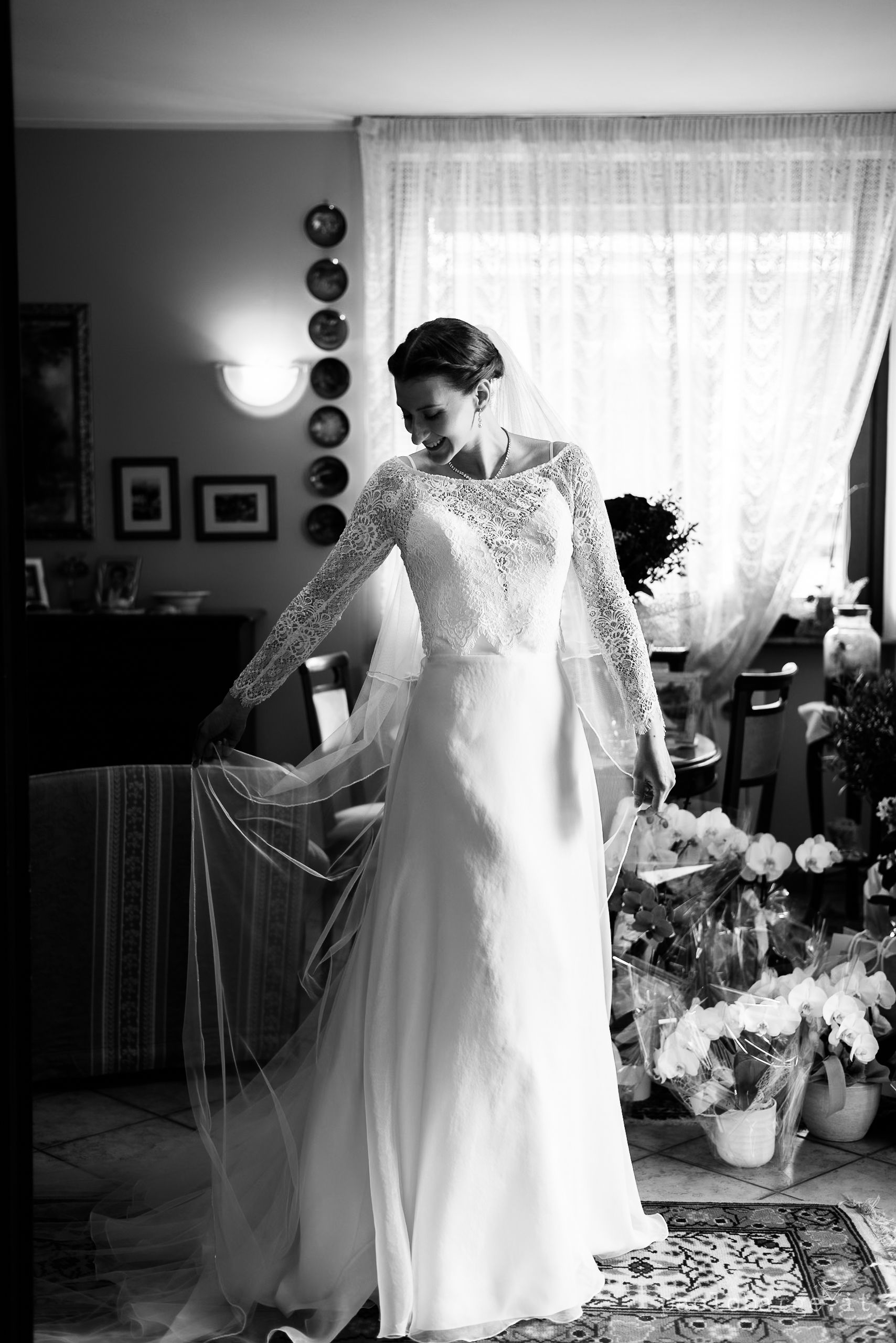 Black and white photograph shows a bride in an elegant lace-sleeve wedding gown admiring her dress in a sunlit room.