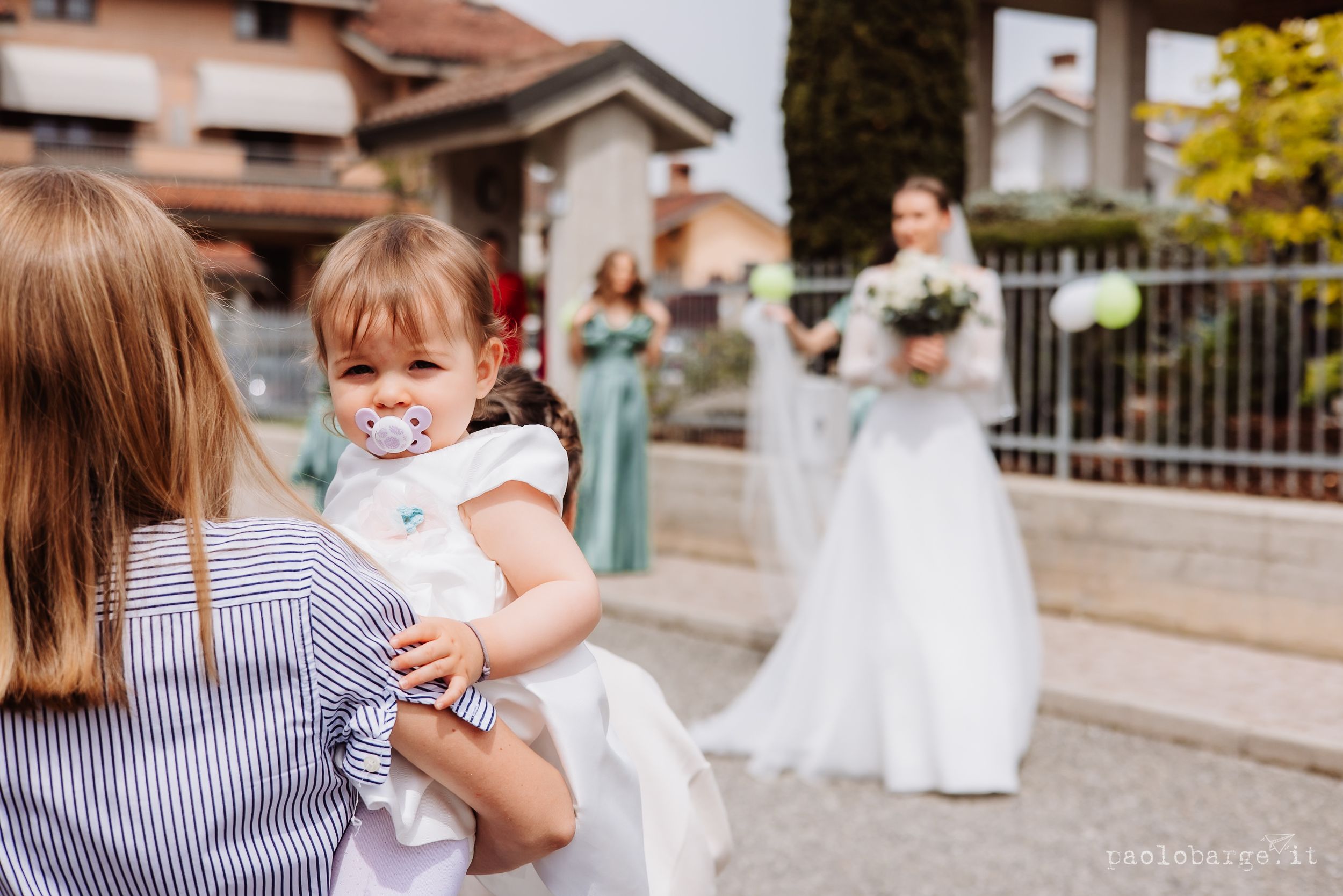 A baby in white watches the bride in a wedding dress in the background while being held by someone in stripes.