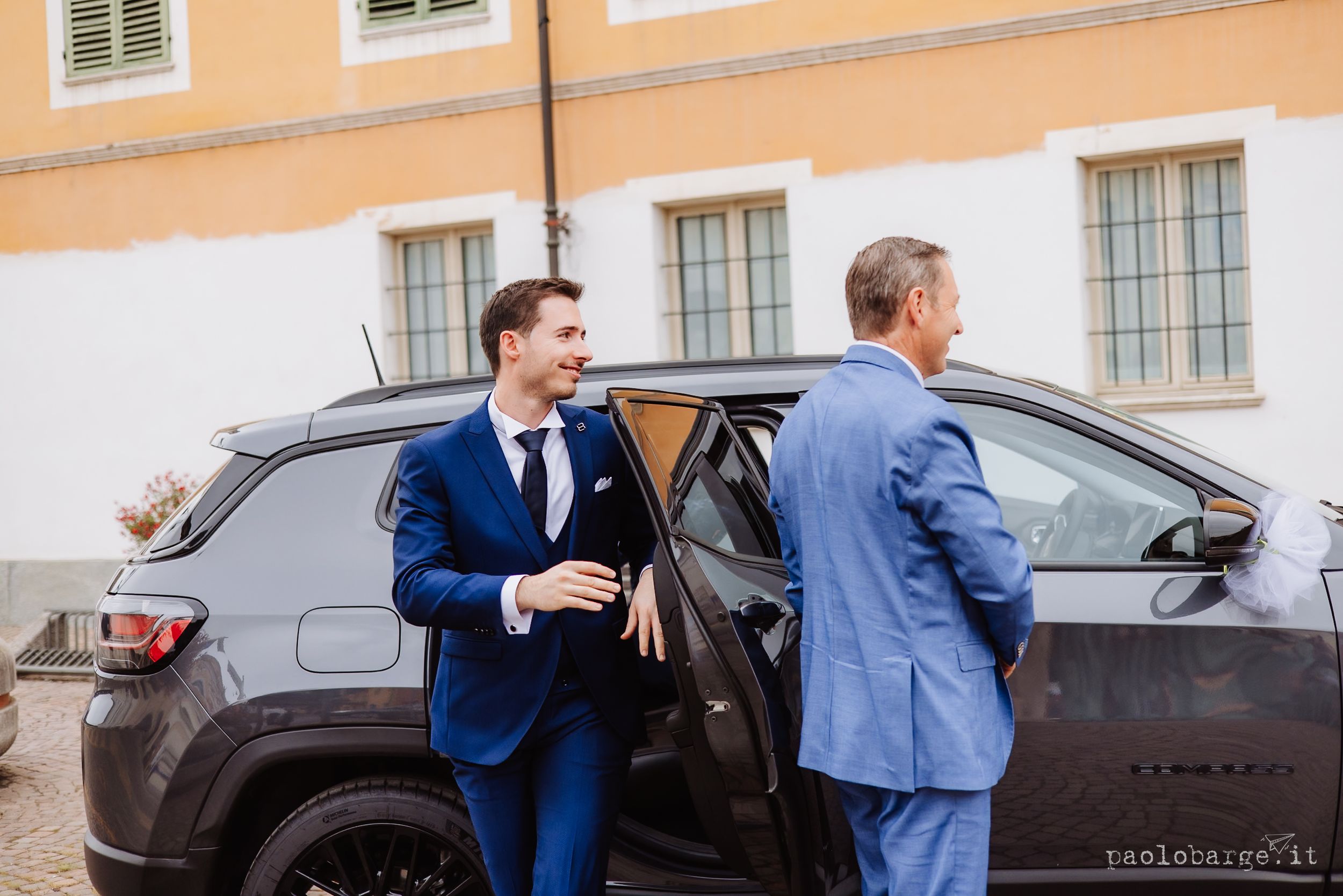People in formal attire interact near luxury vehicles parked outside a building with multiple windows.