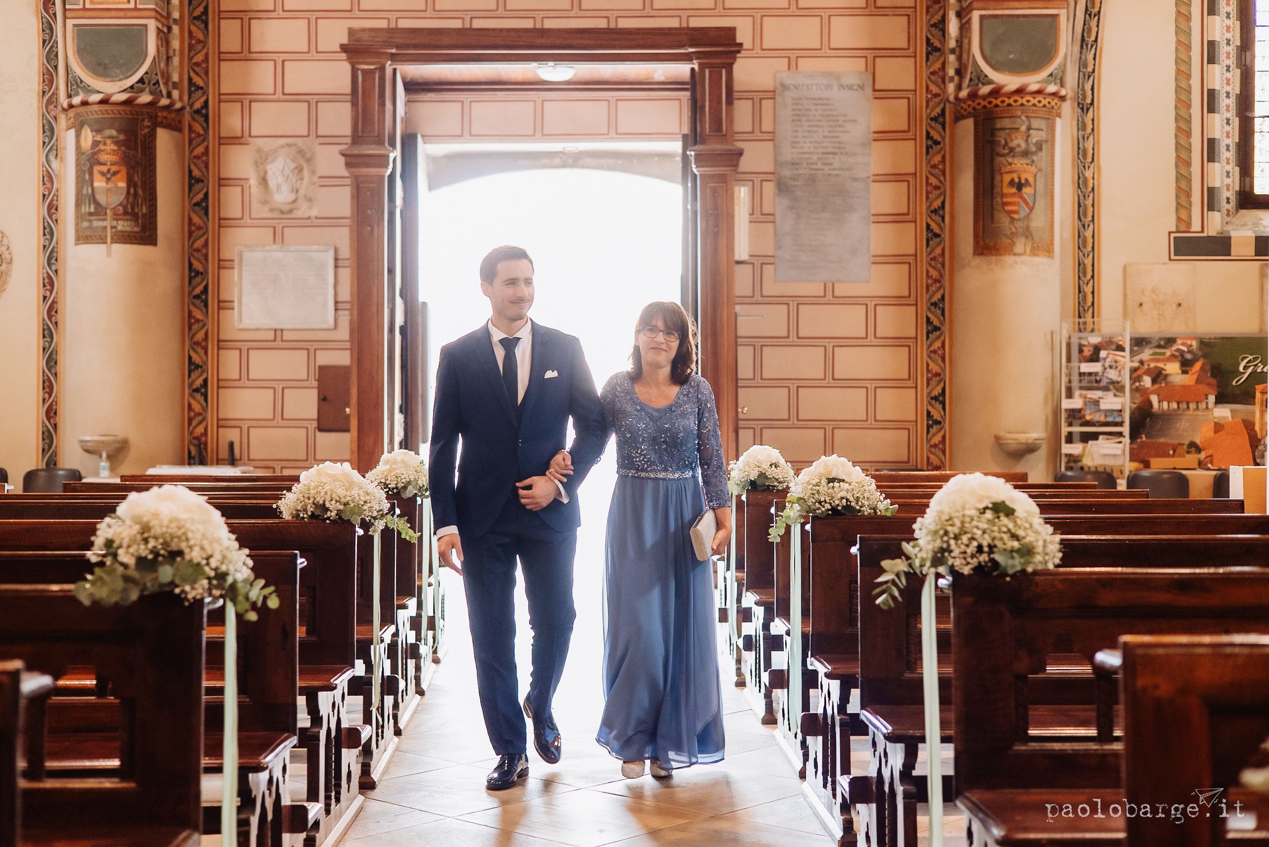 A couple walks down the aisle of a church decorated with white floral arrangements and wooden pews.