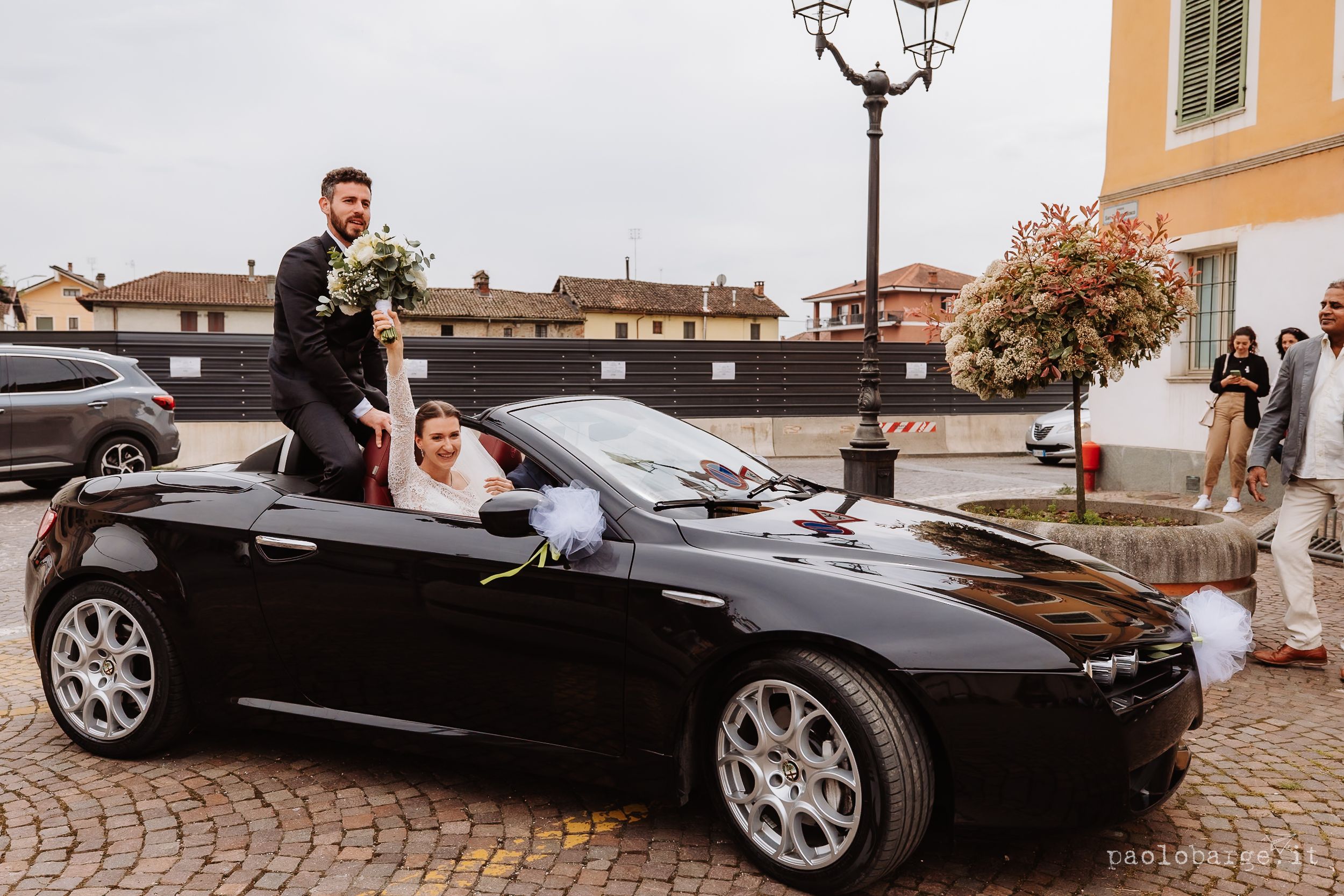 A black convertible sports car parked on cobblestone street during a wedding celebration.