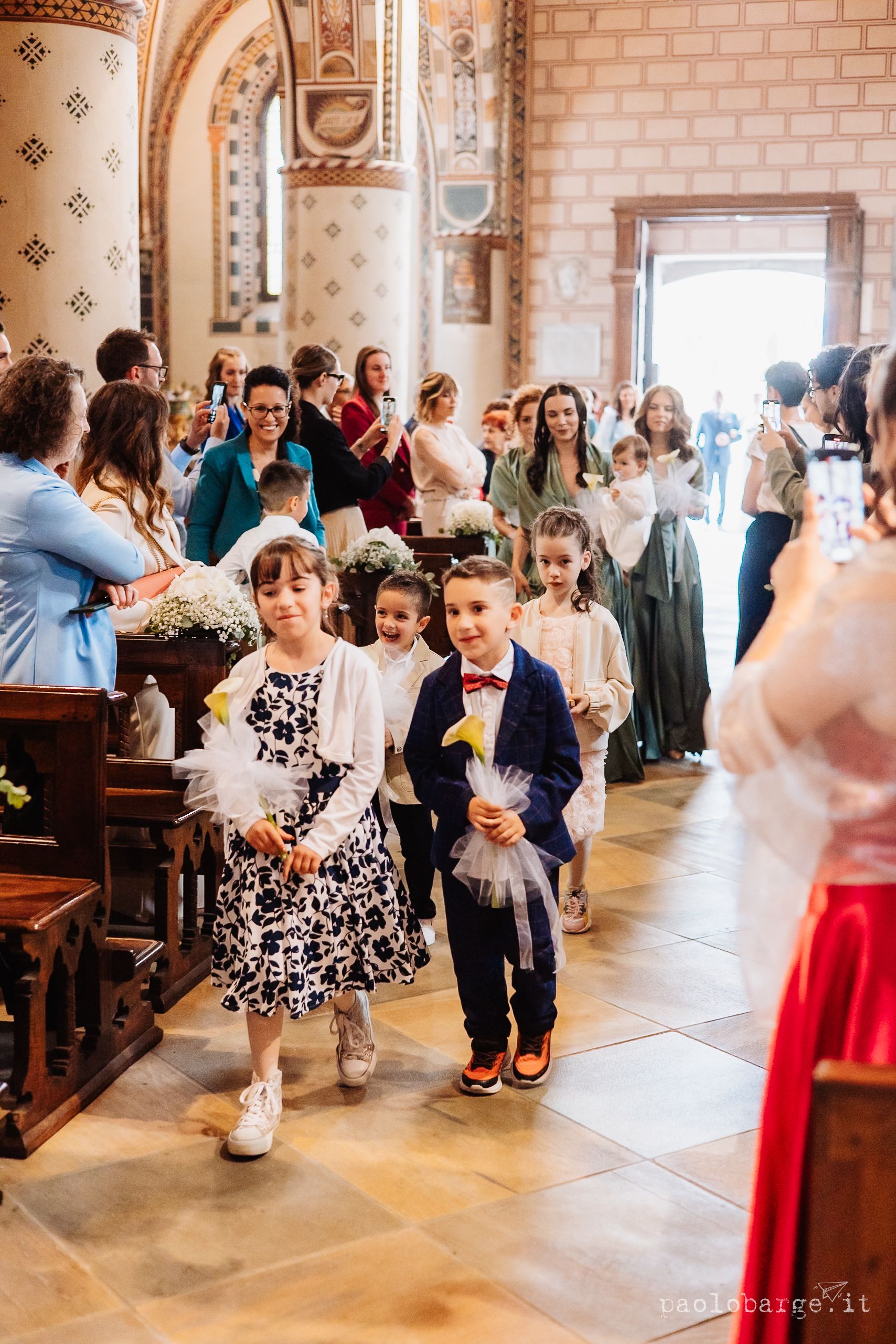 Ring bearers lead wedding guests down the aisle of an ornate church with brick walls and arched details.