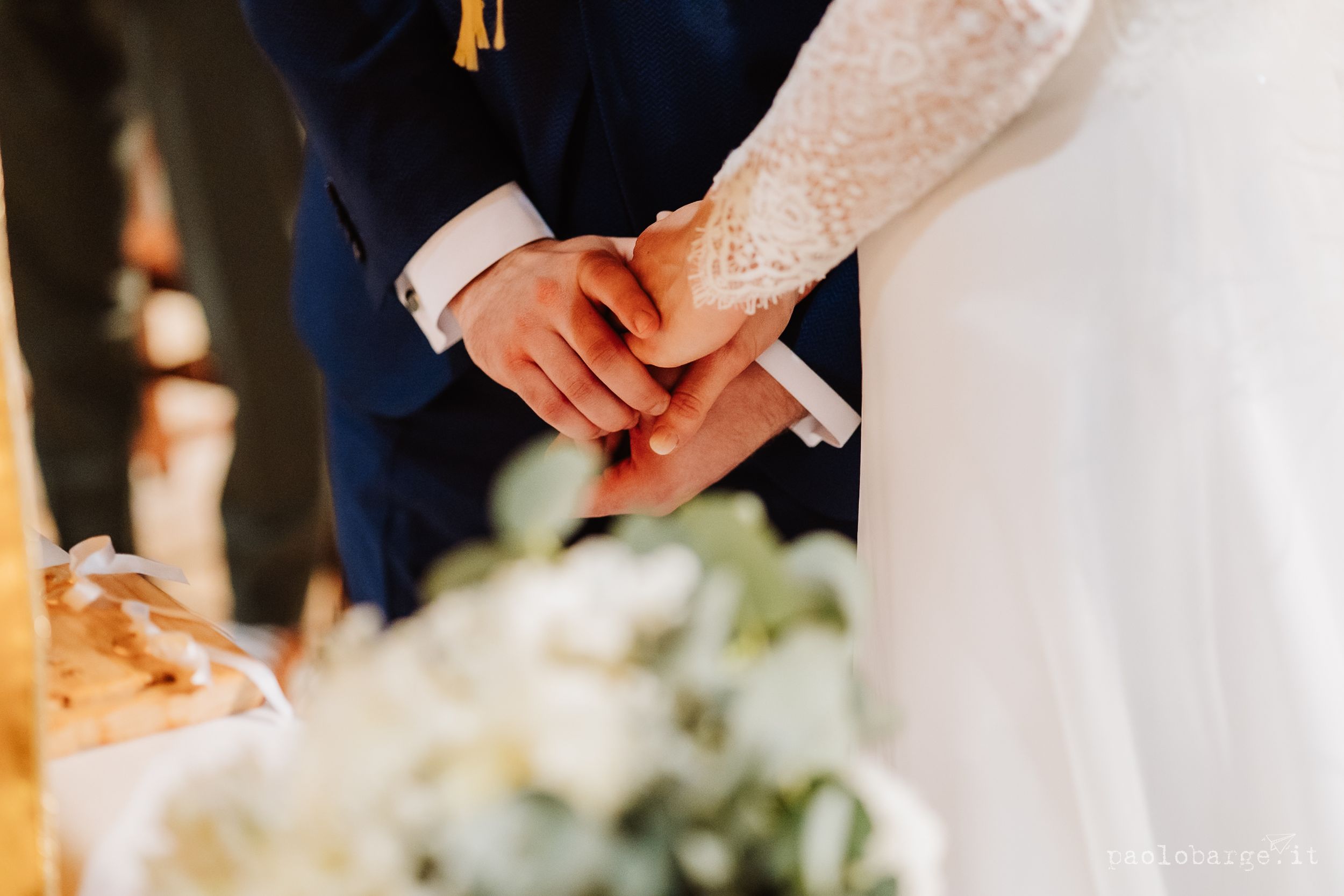 A bride in a white lace gown and groom in a navy suit cut their elegant three-tier wedding cake together.