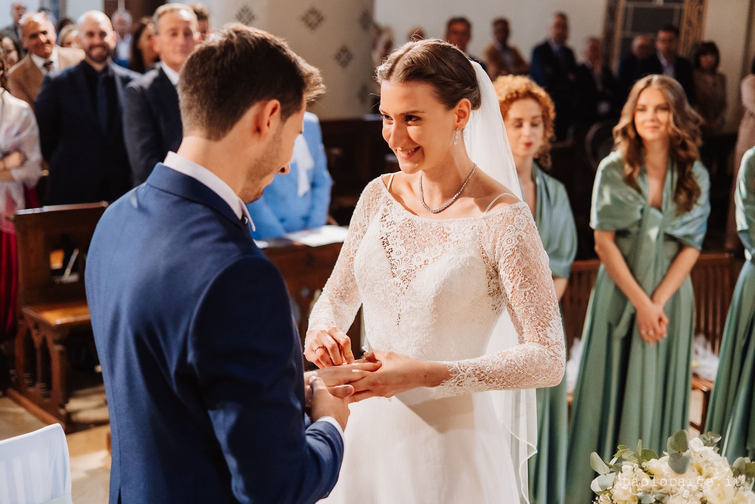 A couple shares an intimate moment while bridesmaids in sage green dresses look on during a wedding ceremony.
