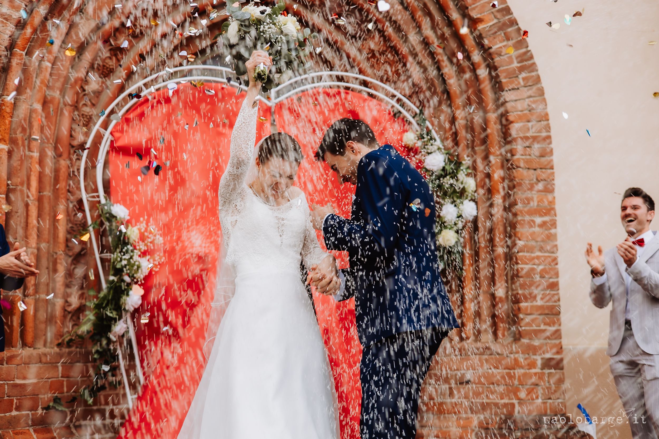 Newlyweds celebrate under a rustic brick arch adorned with flowers as red confetti rains down at an outdoor wedding ceremony.