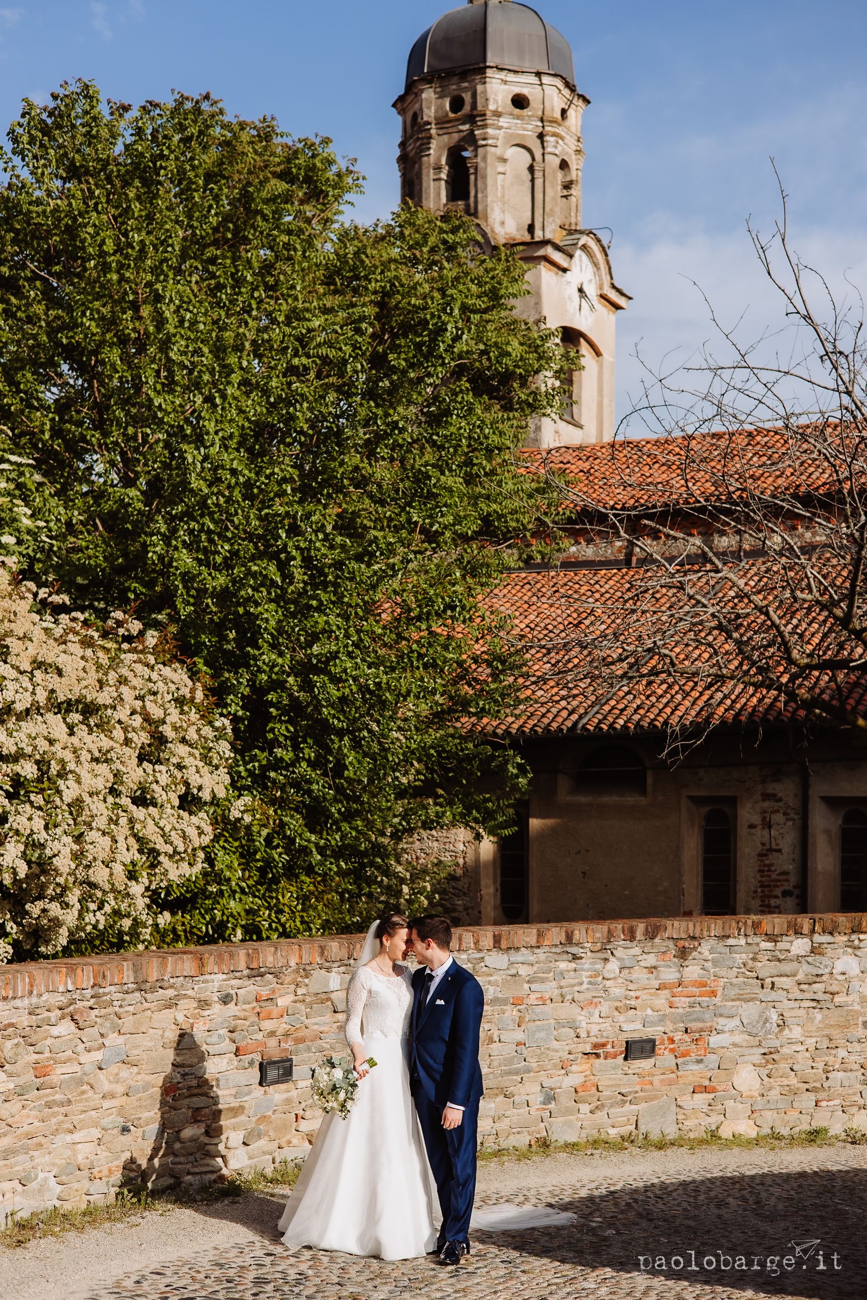 Elegant historic stone church with domed tower and terracotta roof tiles rises behind spring foliage in beautiful Italian setting.