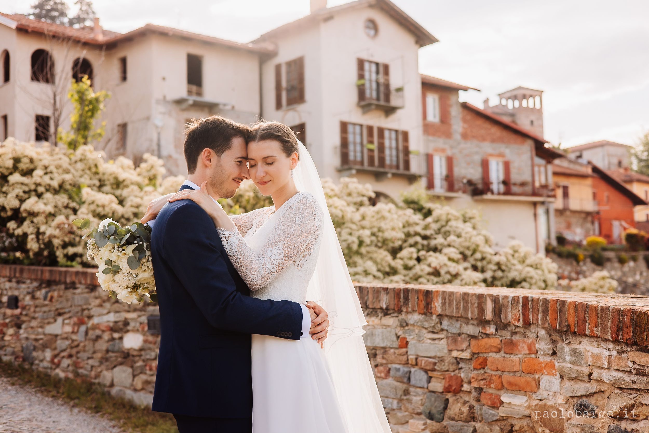 Elegant newlywed couple embraces on stone terrace overlooking Italian village with rustic architecture and blooming flowers.