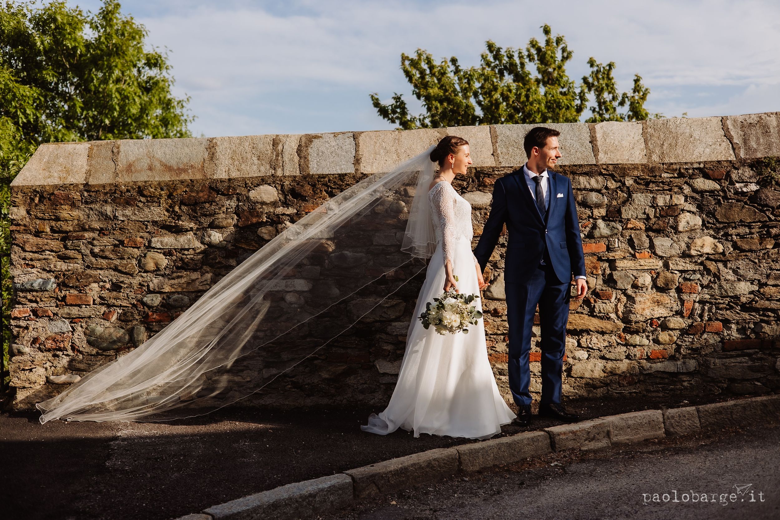 A couple in wedding attire stands together near a rustic stone wall as a long veil catches the sunlight.
