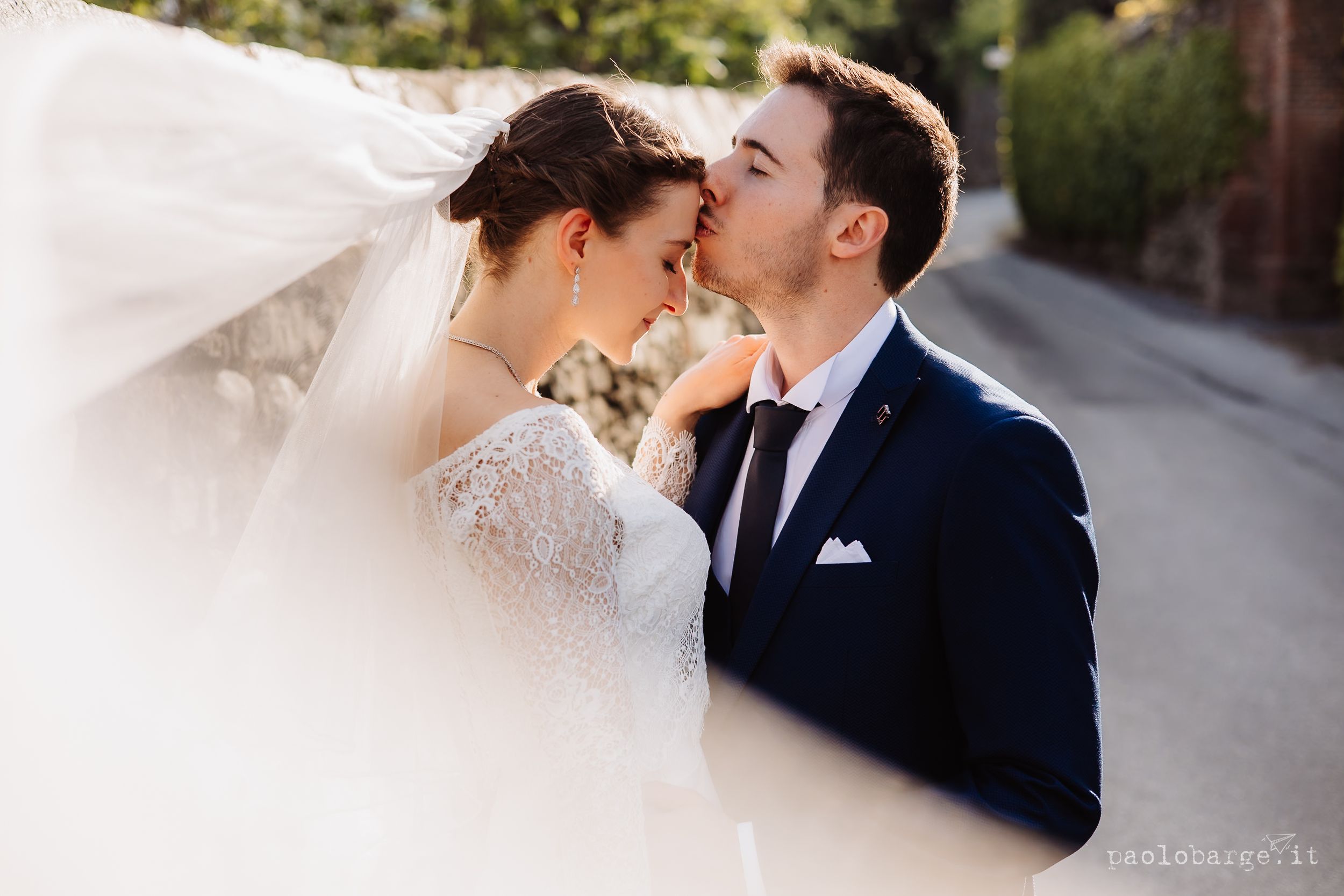 A romantic wedding photo showing a bride in white lace and groom in navy suit sharing a tender moment outdoors.