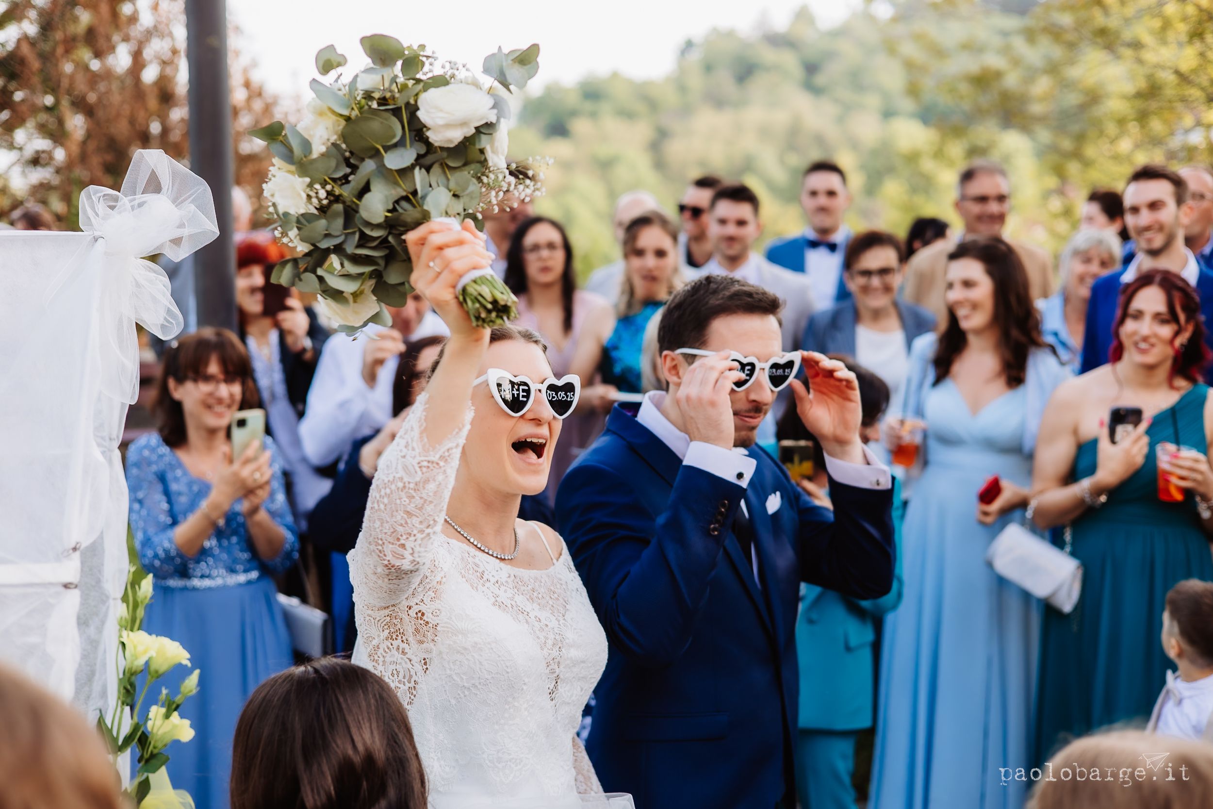 Wedding guests celebrate at outdoor reception with guests in blue dresses and dark suits during daytime celebration.