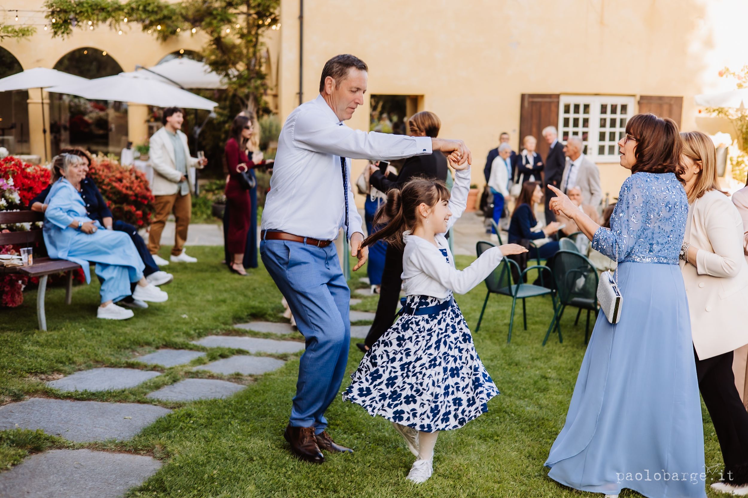 A man in blue shirt and young girl in a patterned dress dance together at an outdoor garden party event.