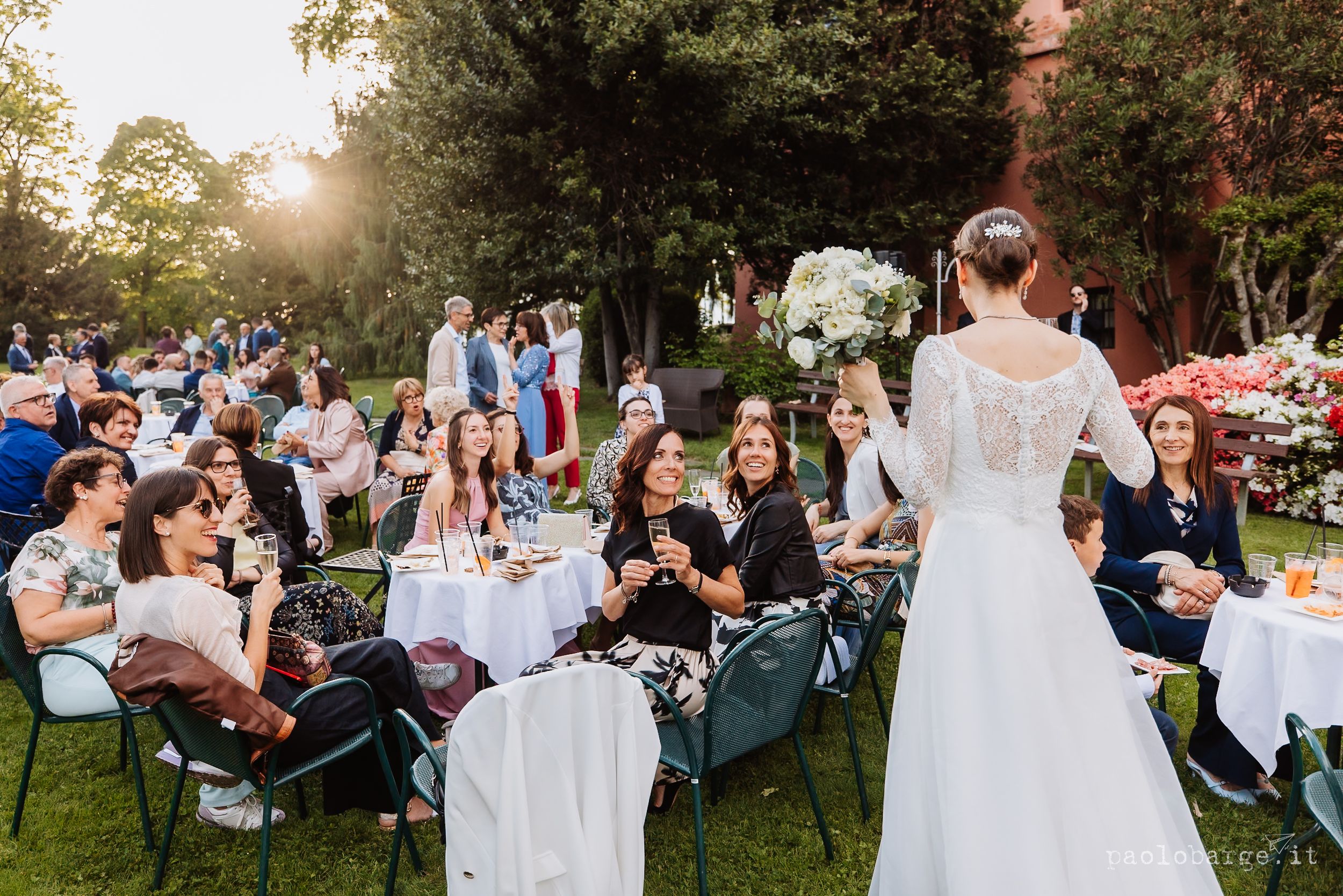 Wedding guests sit at outdoor garden reception tables while a person in a white dress stands at one end during sunset.