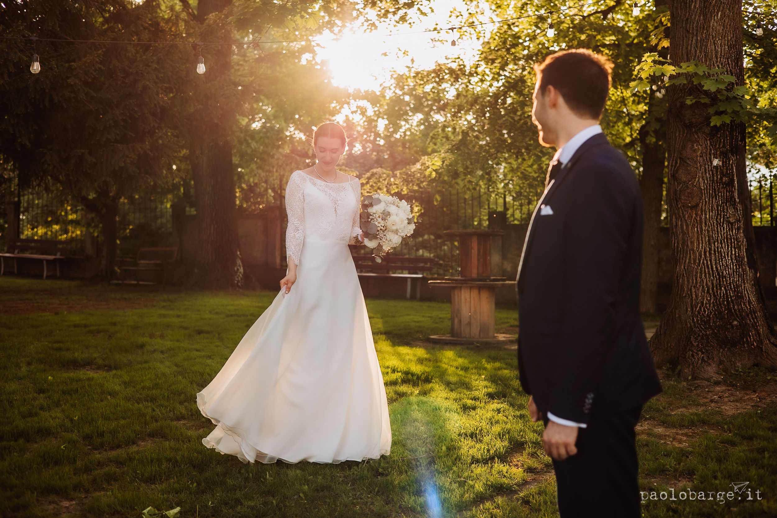 A romantic wedding couple stands in a sunlit garden as golden light streams through the trees at sunset.