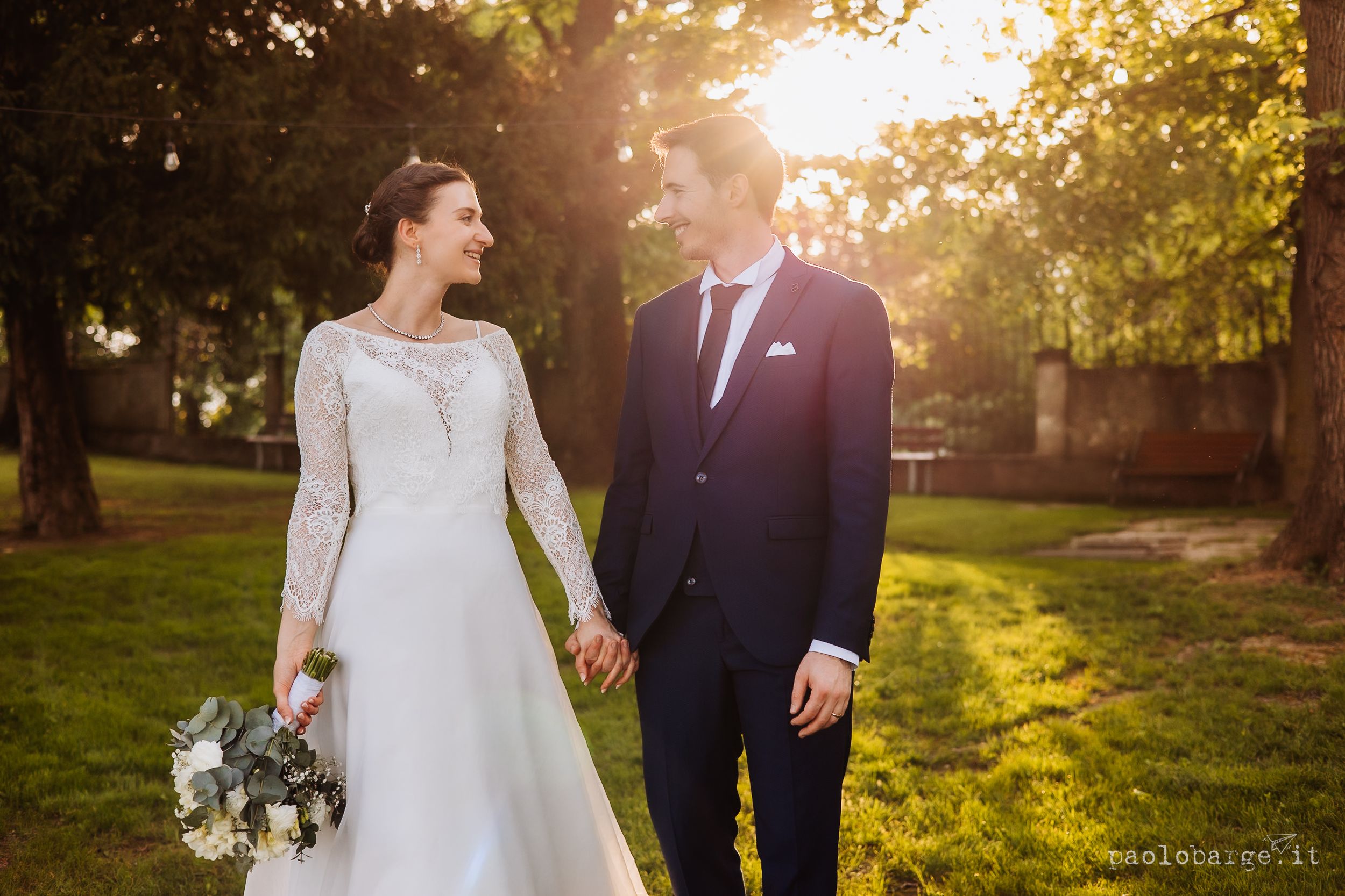 A couple holding hands while walking in a sunlit park during golden hour, with the bride wearing a long-sleeved white gown.