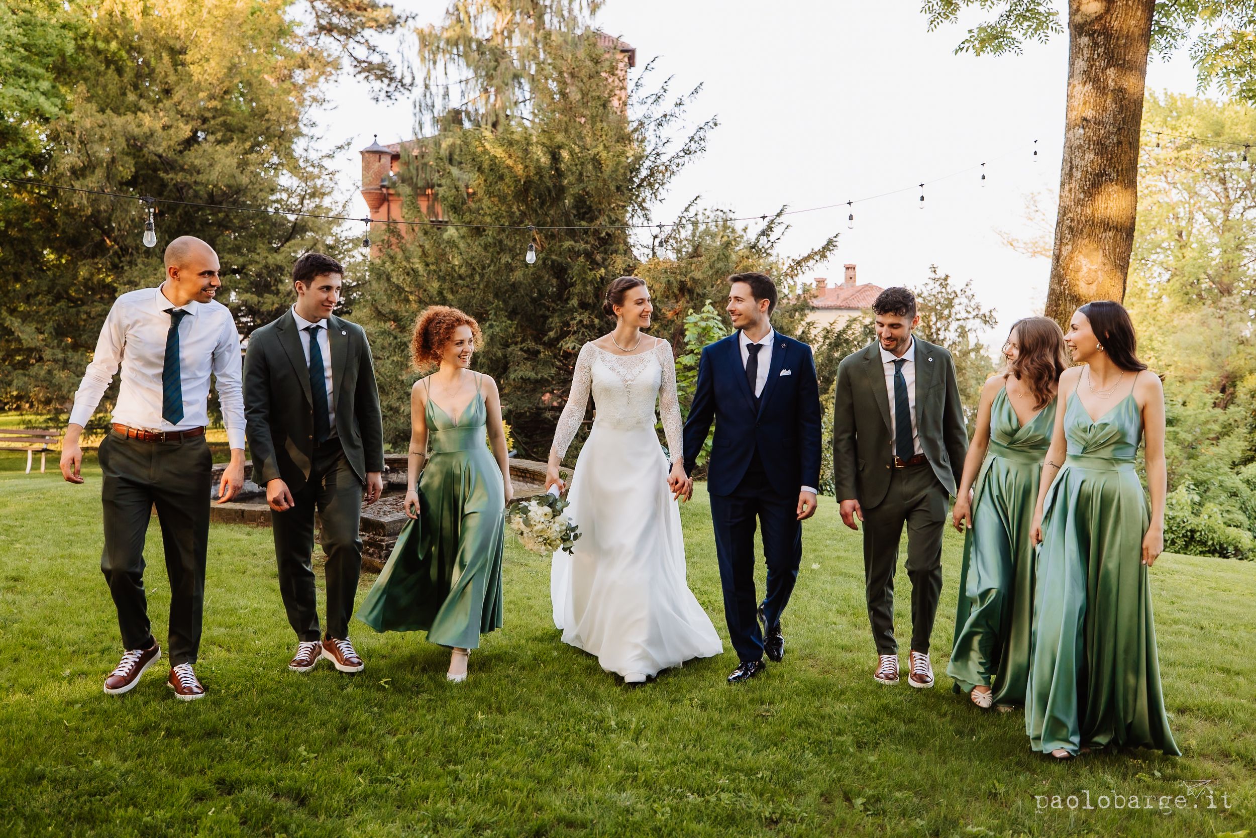 Wedding party walks together on grass lawn wearing mint green bridesmaid dresses and dark suits for outdoor celebration.