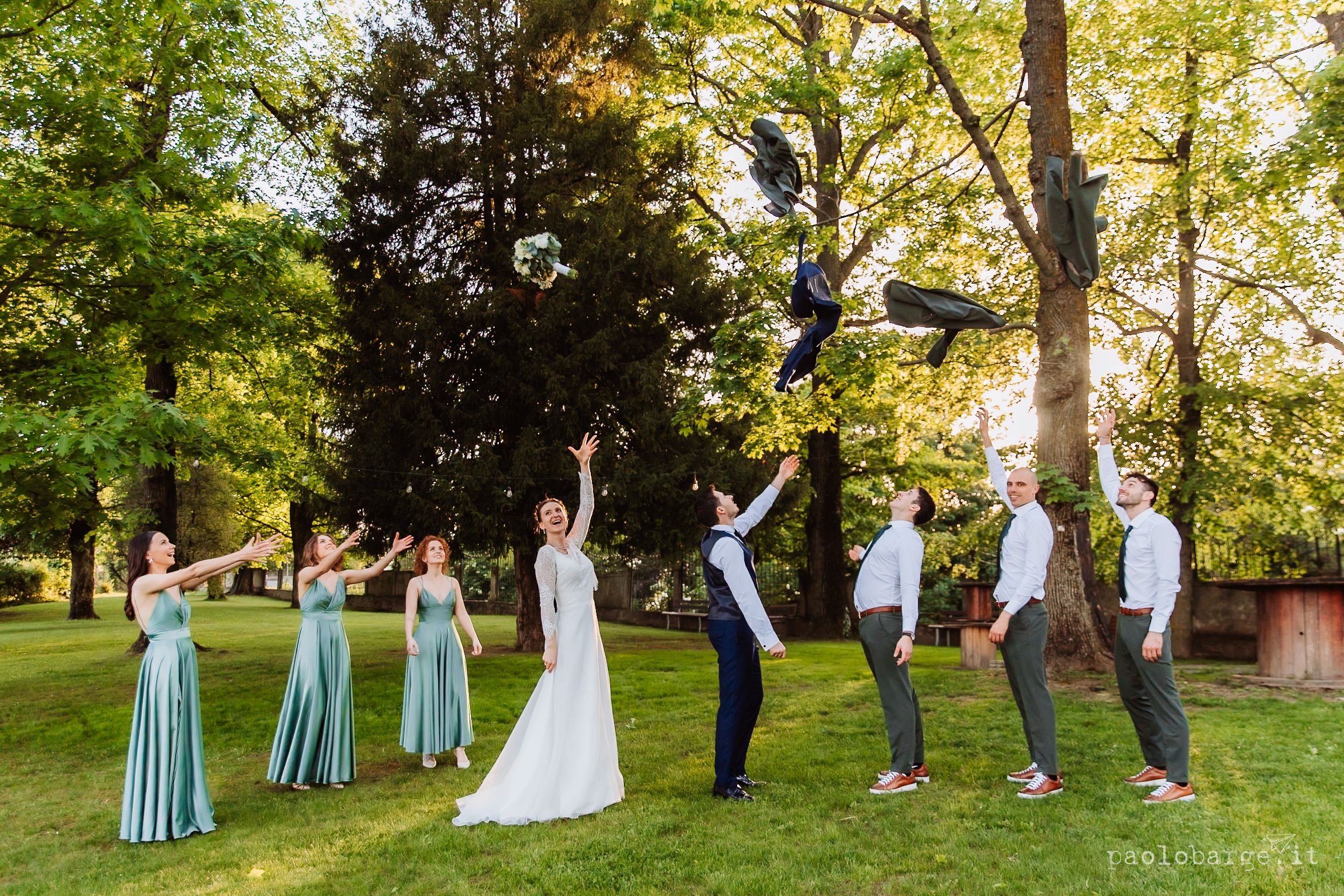 Wedding party strikes fun poses in a line on a lush green lawn during golden hour photography session.