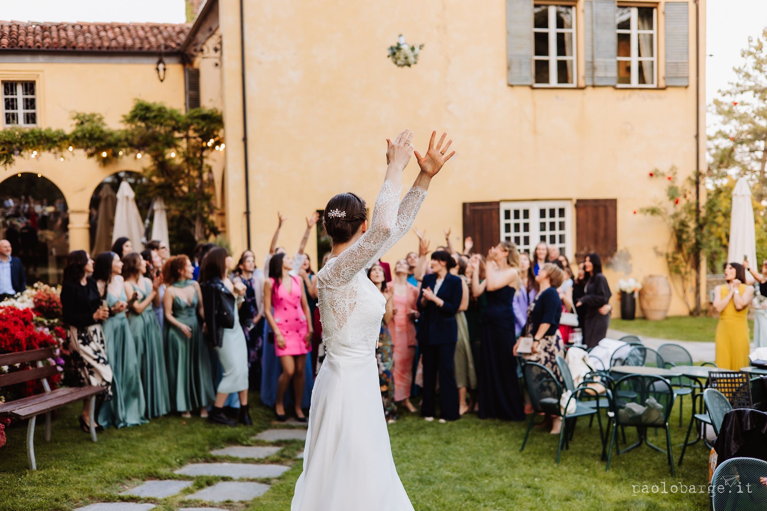 Bride tosses her bouquet to a crowd of wedding guests gathered outside a Mediterranean-style villa.