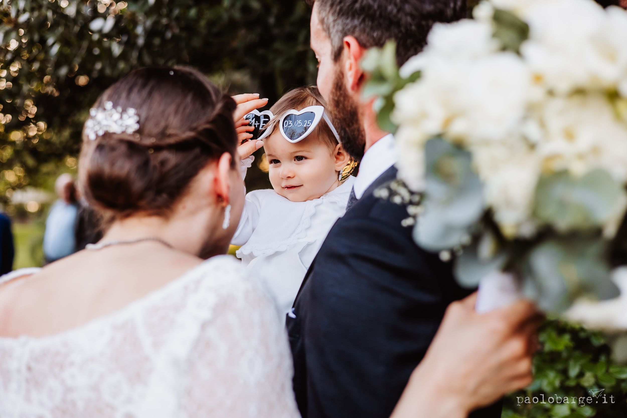 A small child wearing sunglasses peeks between adults in formal wedding attire holding white flowers.