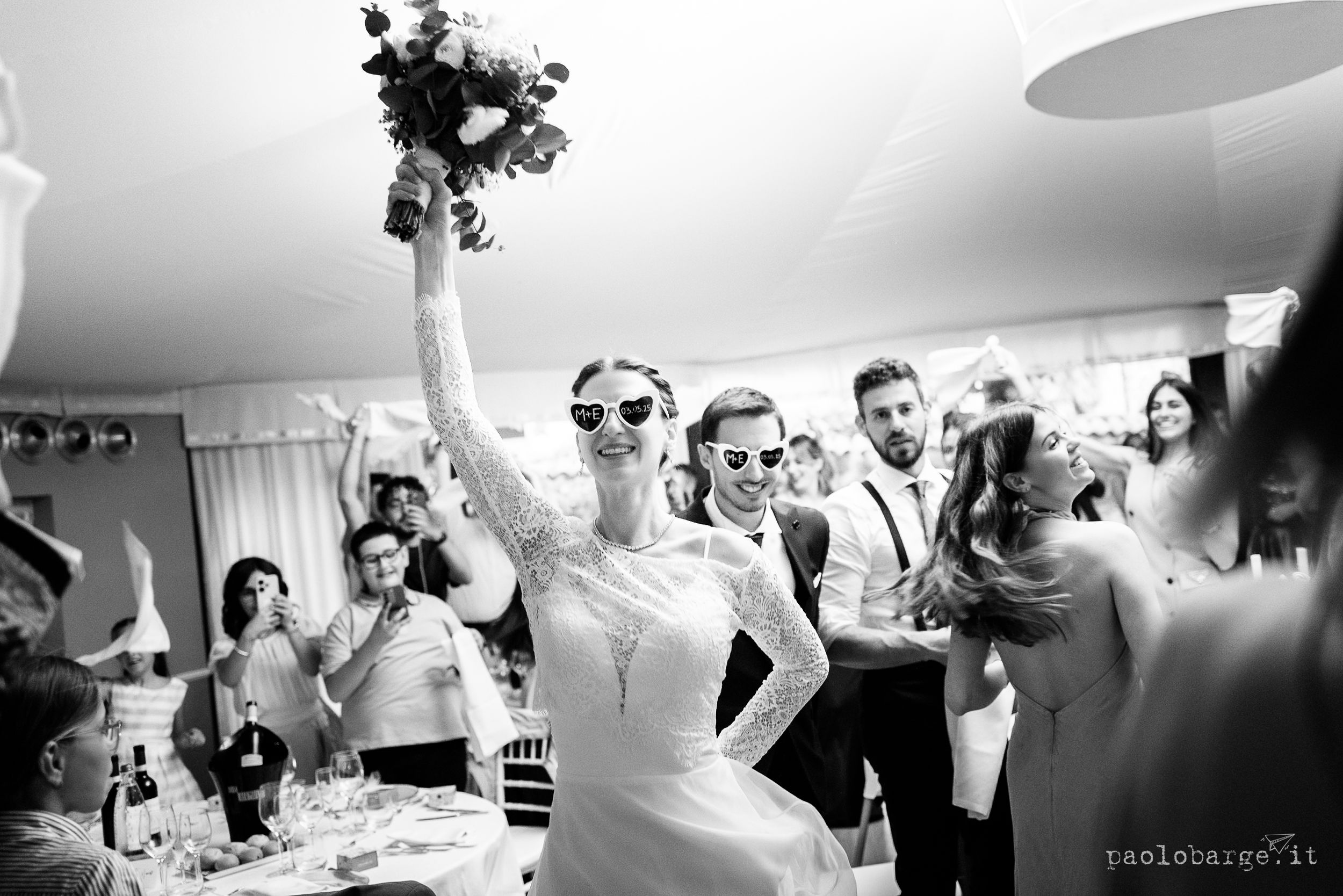 A joyful wedding party celebrates during a reception as the bride energetically raises her bouquet in a black and white photo.