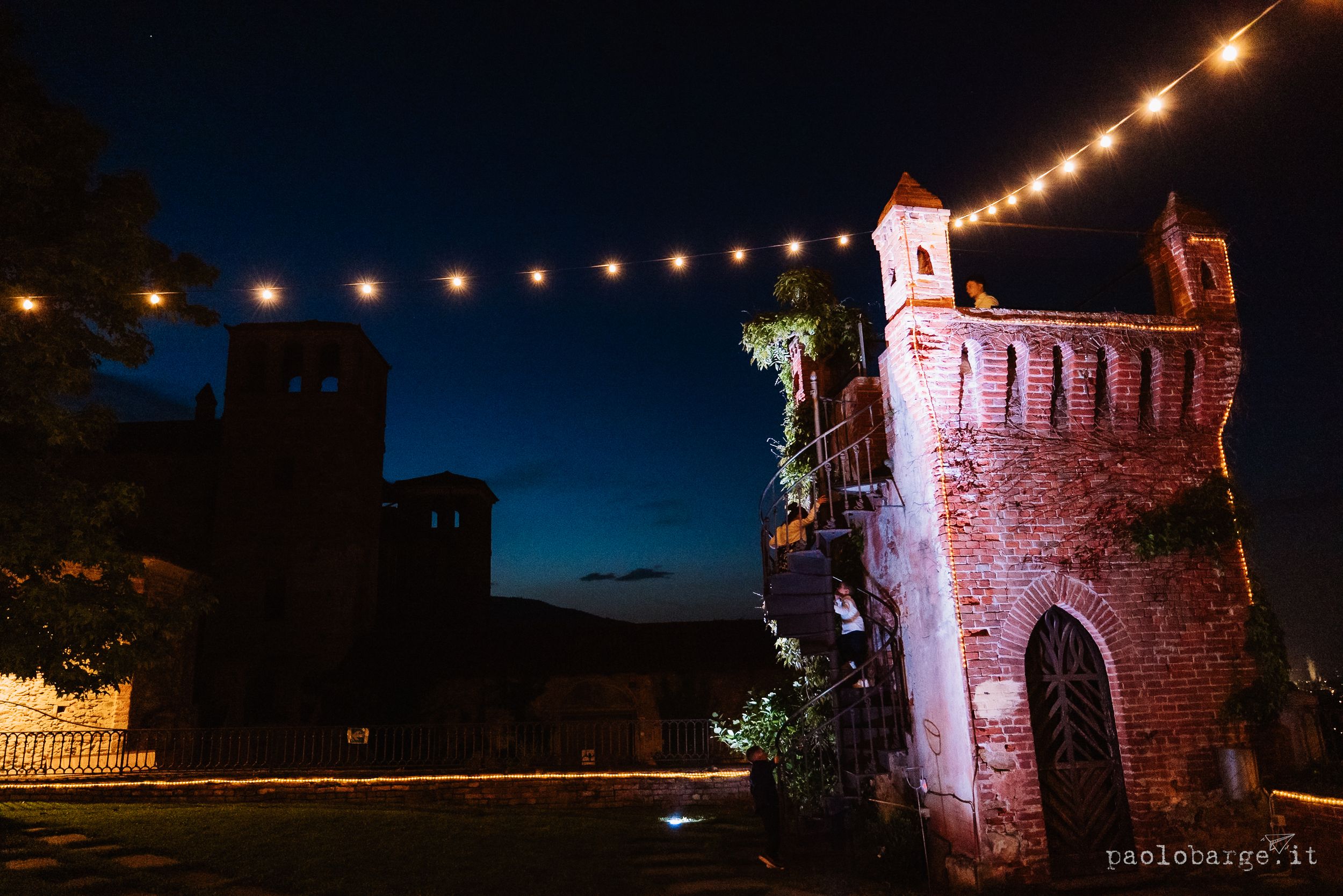 Ancient stone castle tower illuminated in pink at night with string lights creating a romantic ambiance against dark sky.
